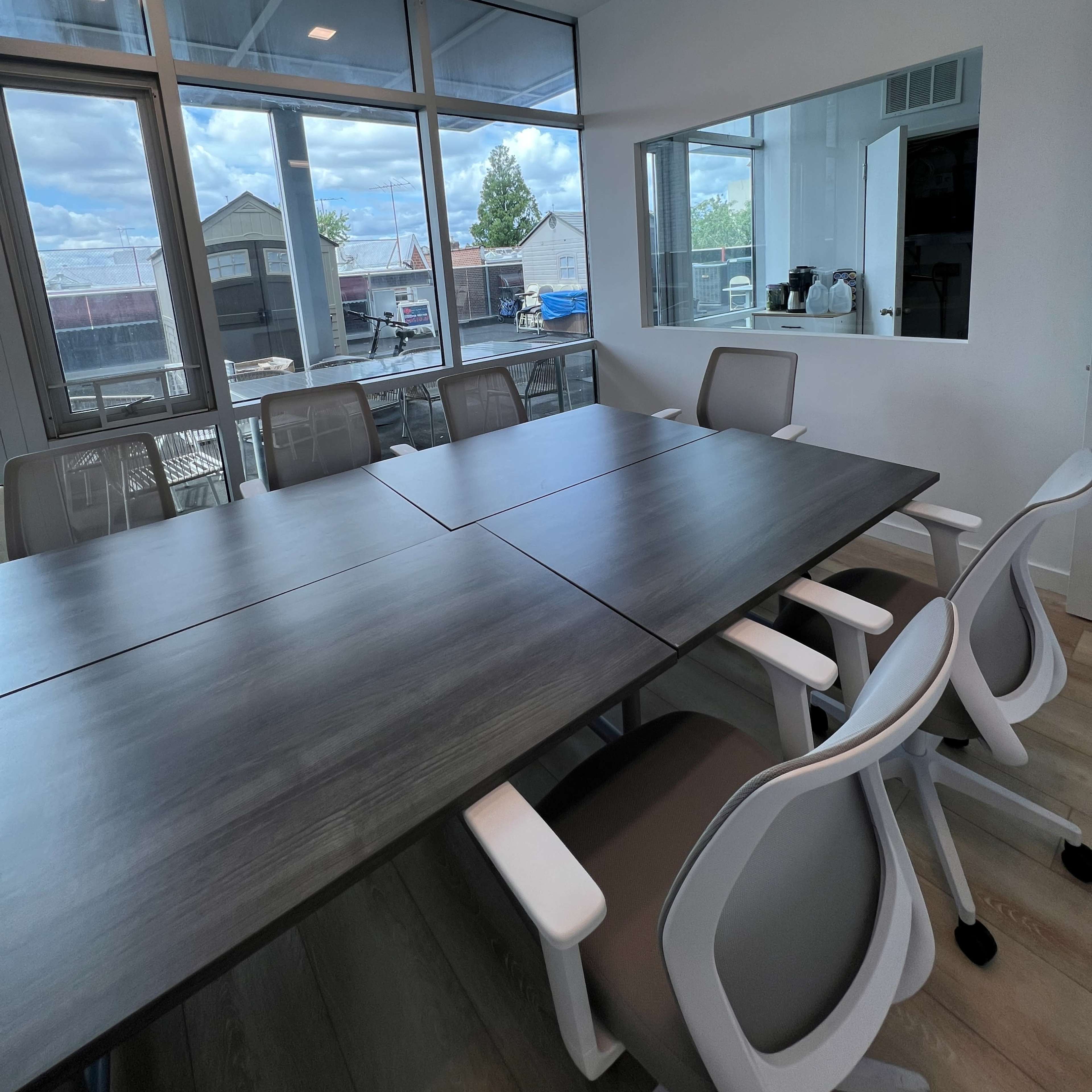 A modern conference room features a large table surrounded by white chairs and large windows overlooking an outdoor area.