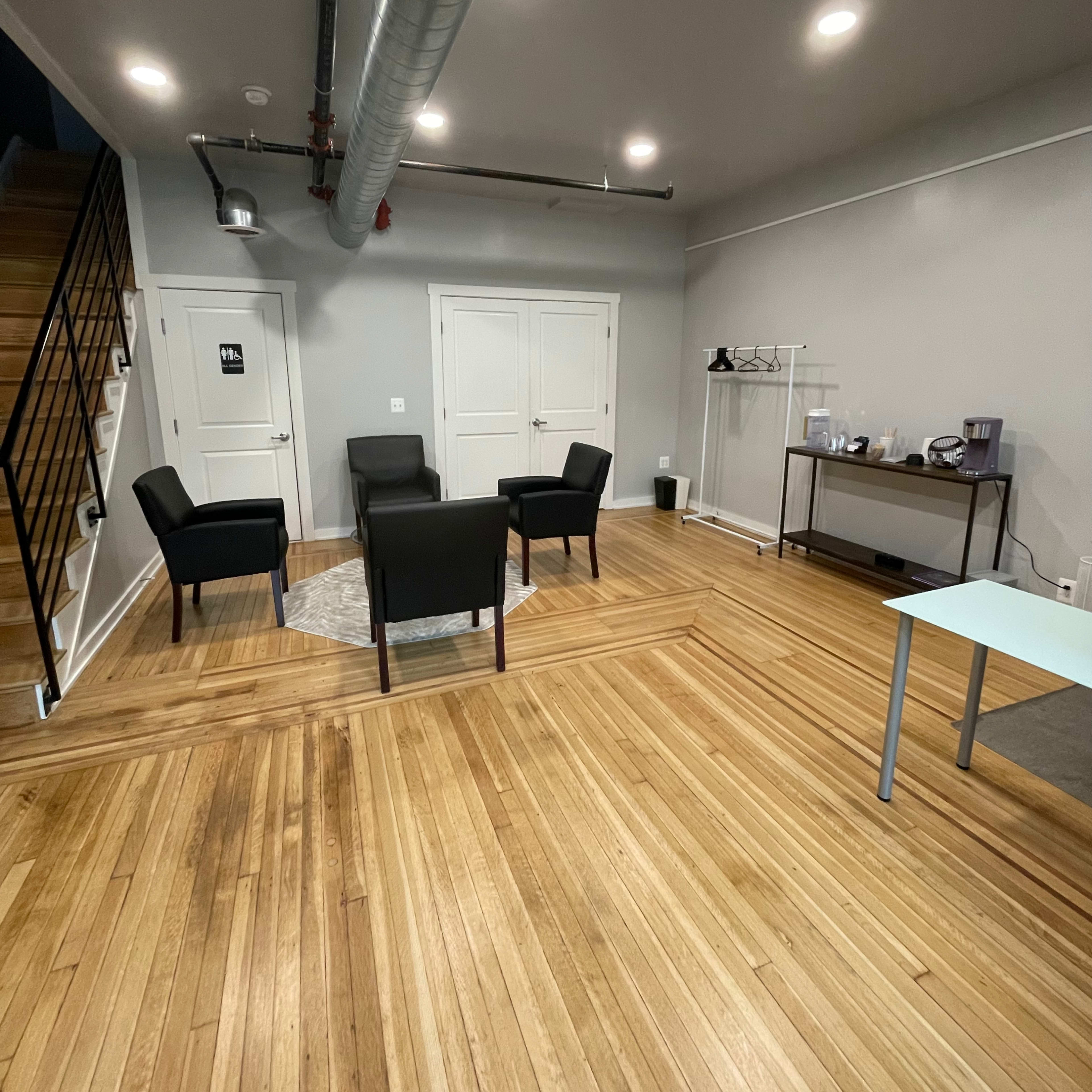 A modern interior space with wooden flooring, featuring a seating area with four black chairs around a circular rug and a small table against a wall.