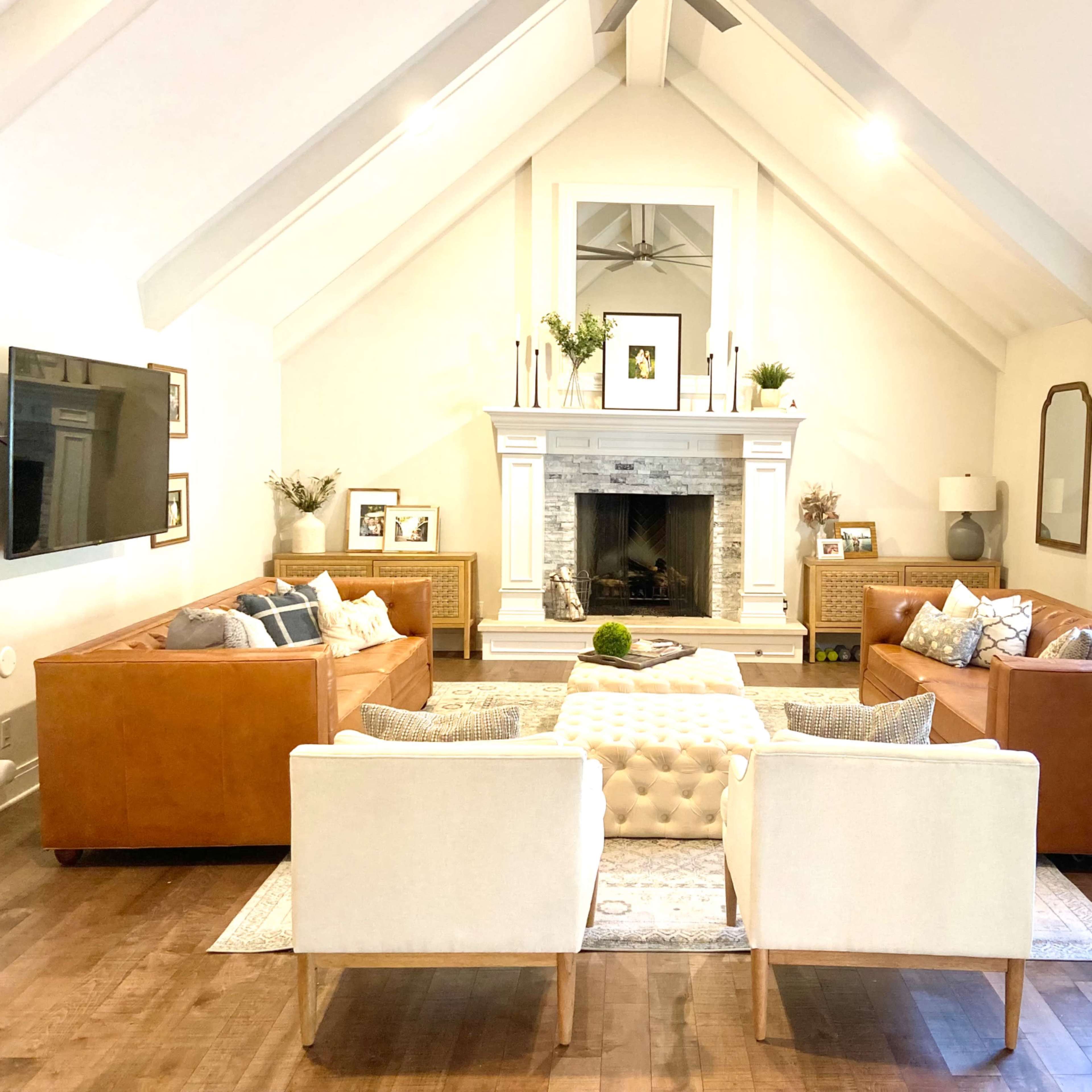 A spacious living room features tan leather sofas and white armchairs arranged around a central tufted coffee table in front of a stone fireplace.