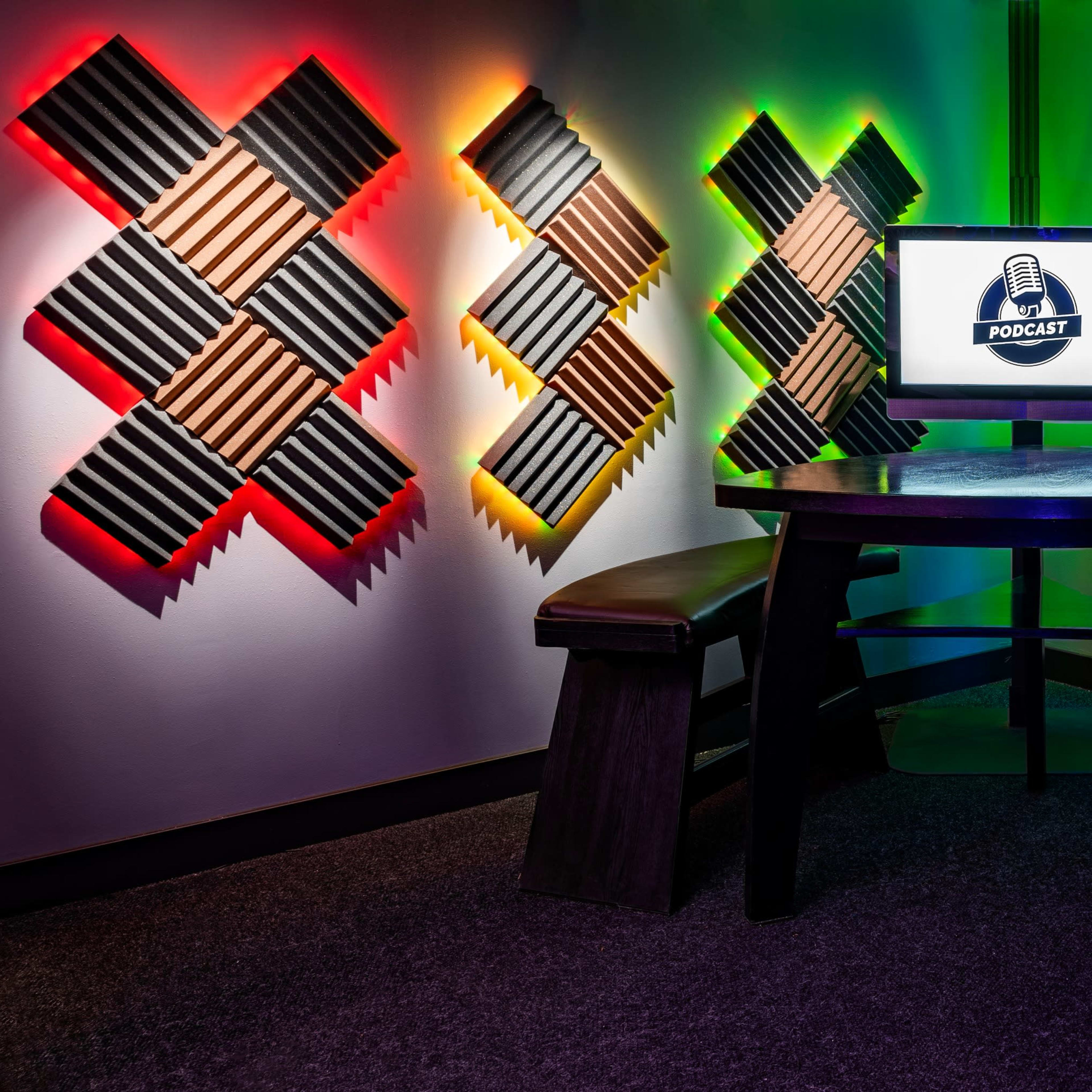 A podcasting studio features a central table with two benches, illuminated by colorful LED lights and surrounded by soundproofing panels.