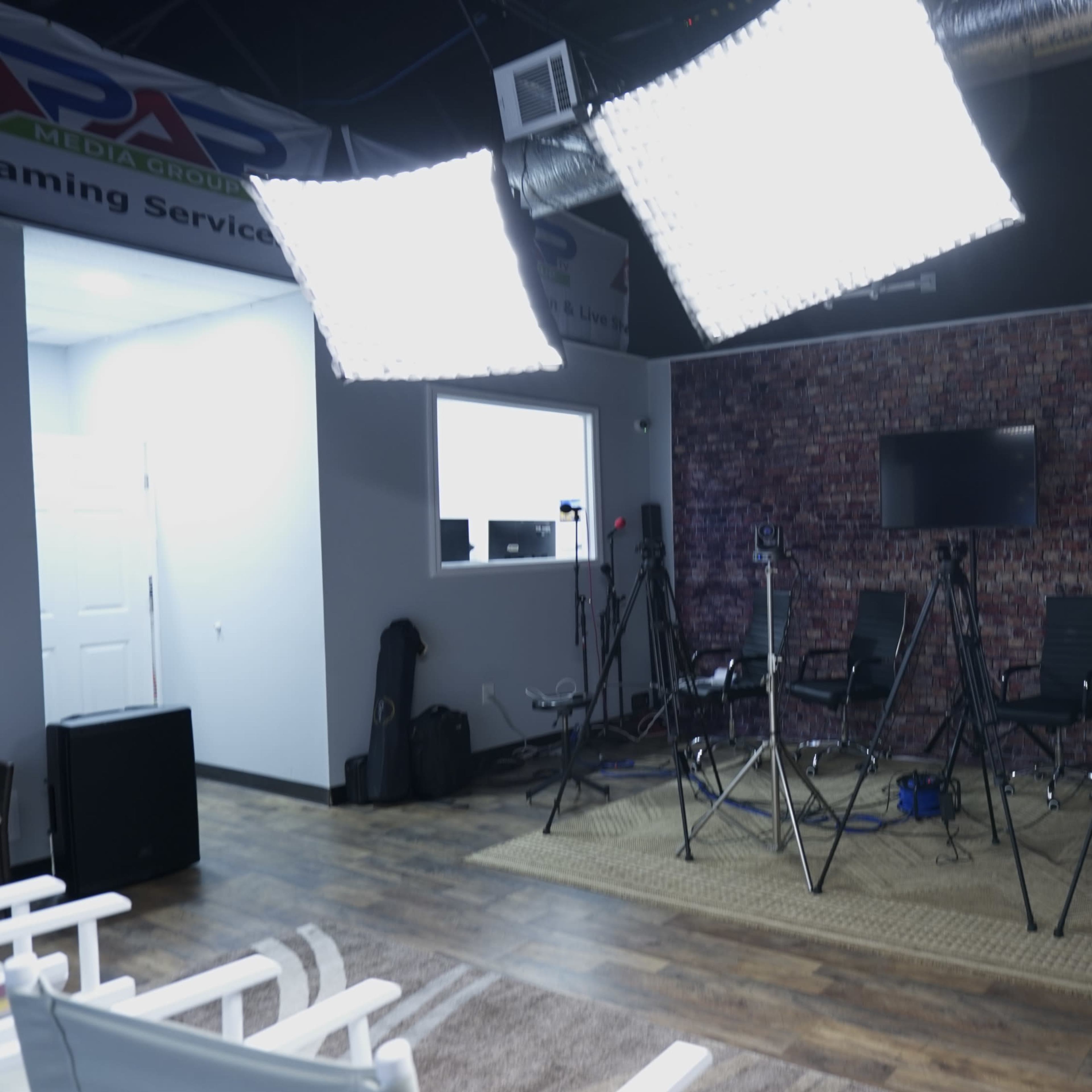 A studio setup features multiple camera tripods, lighting equipment, and a green screen against a brick wall backdrop.