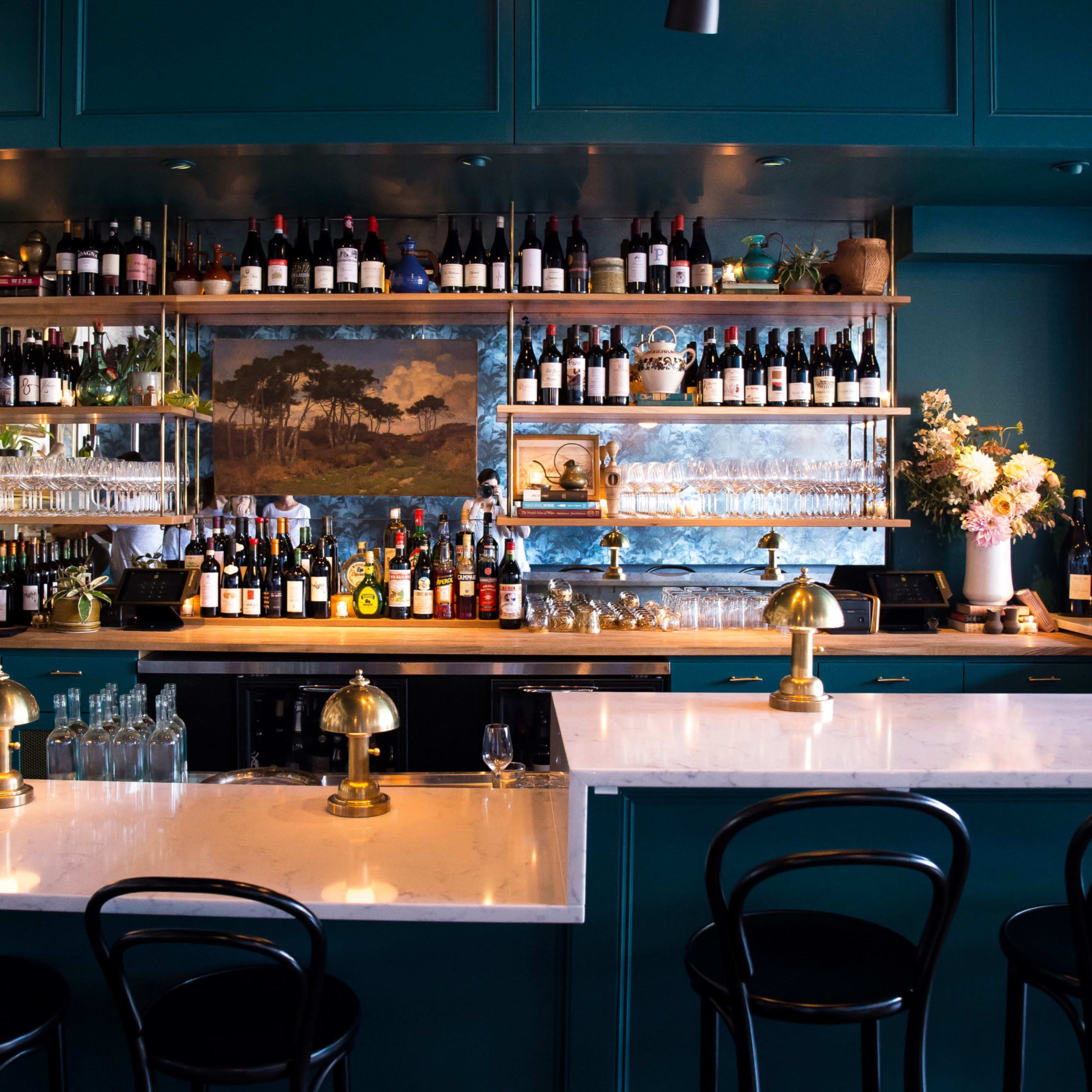 A stylish bar with teal cabinetry, shelves filled with an array of wine bottles, and two marble-topped counters with brass fixtures.