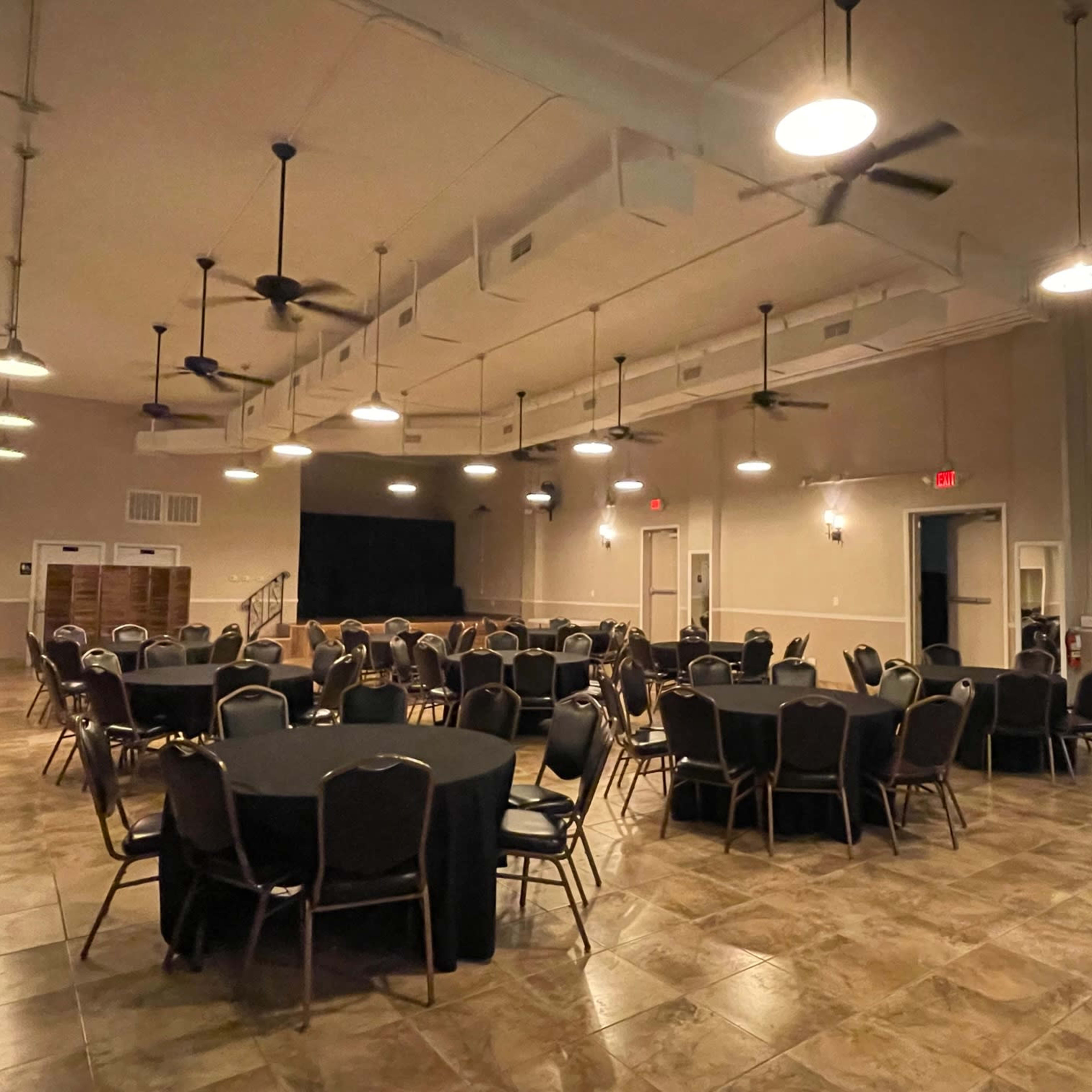 A large indoor event space features several round tables covered with black tablecloths, arranged in a grid pattern beneath ceiling fans and overhead lights.