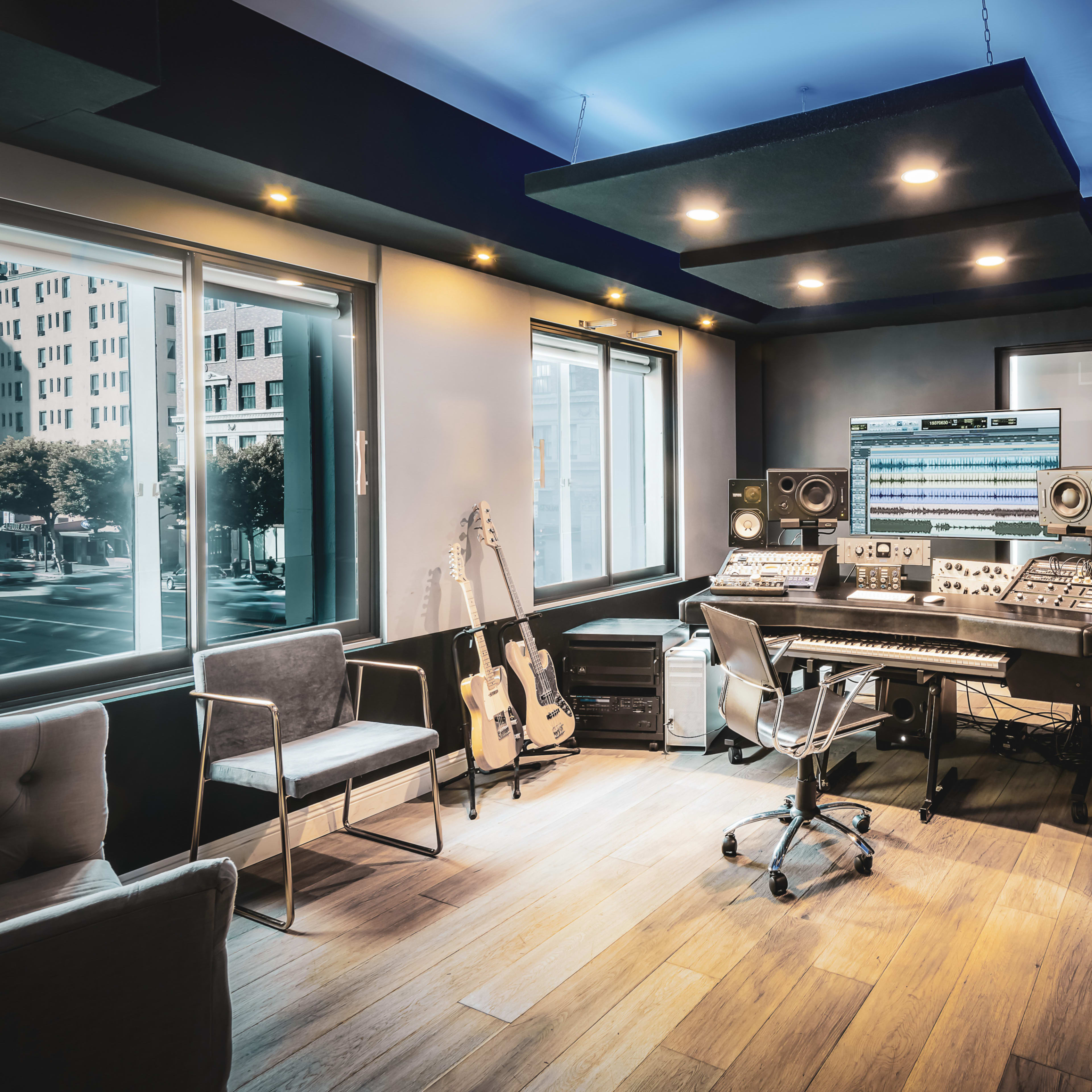 A modern music studio features a mix of acoustic panels, recording equipment, and musical instruments, with large windows allowing natural light.