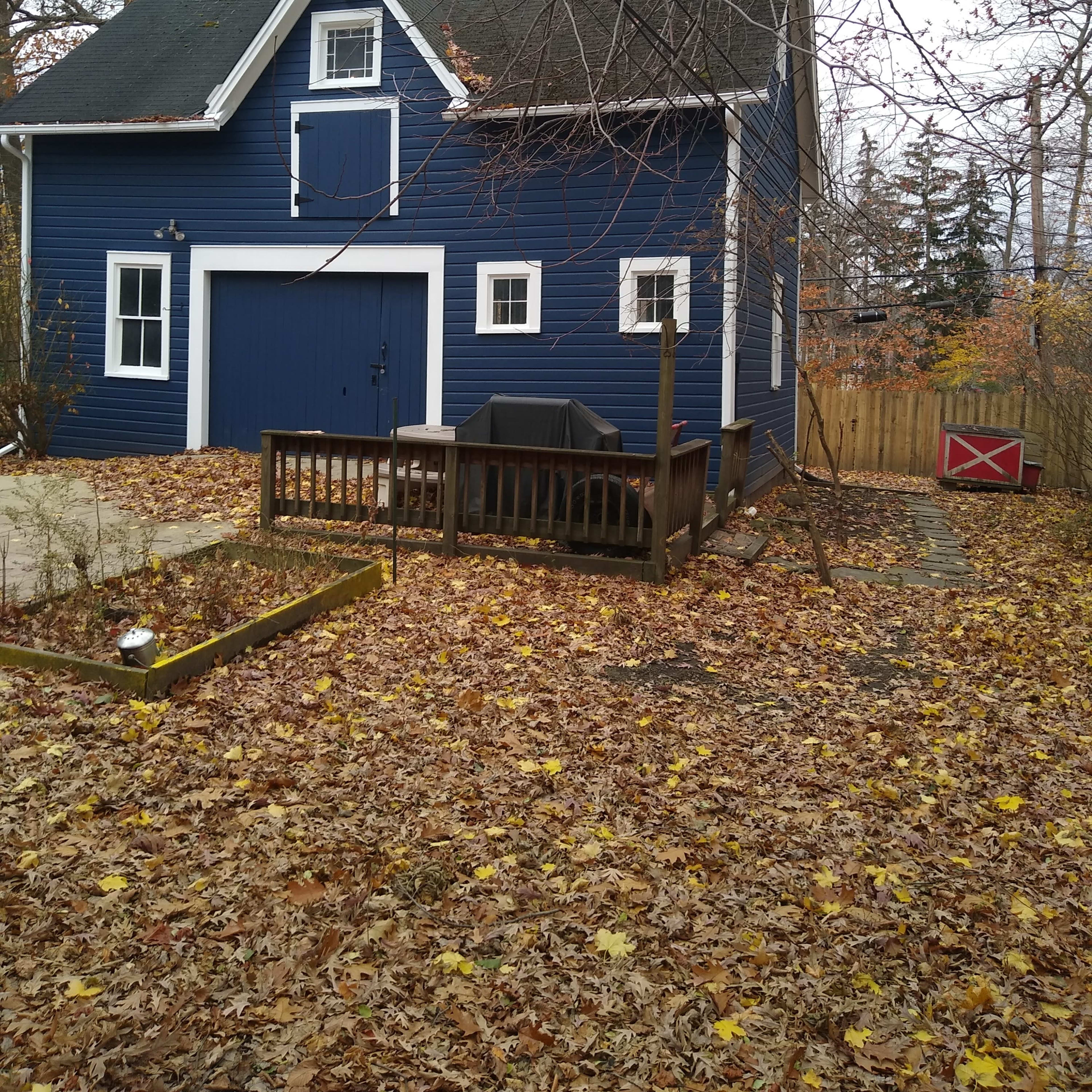 A blue two-story house with a deck, surrounded by fallen leaves and trees in a suburban setting.