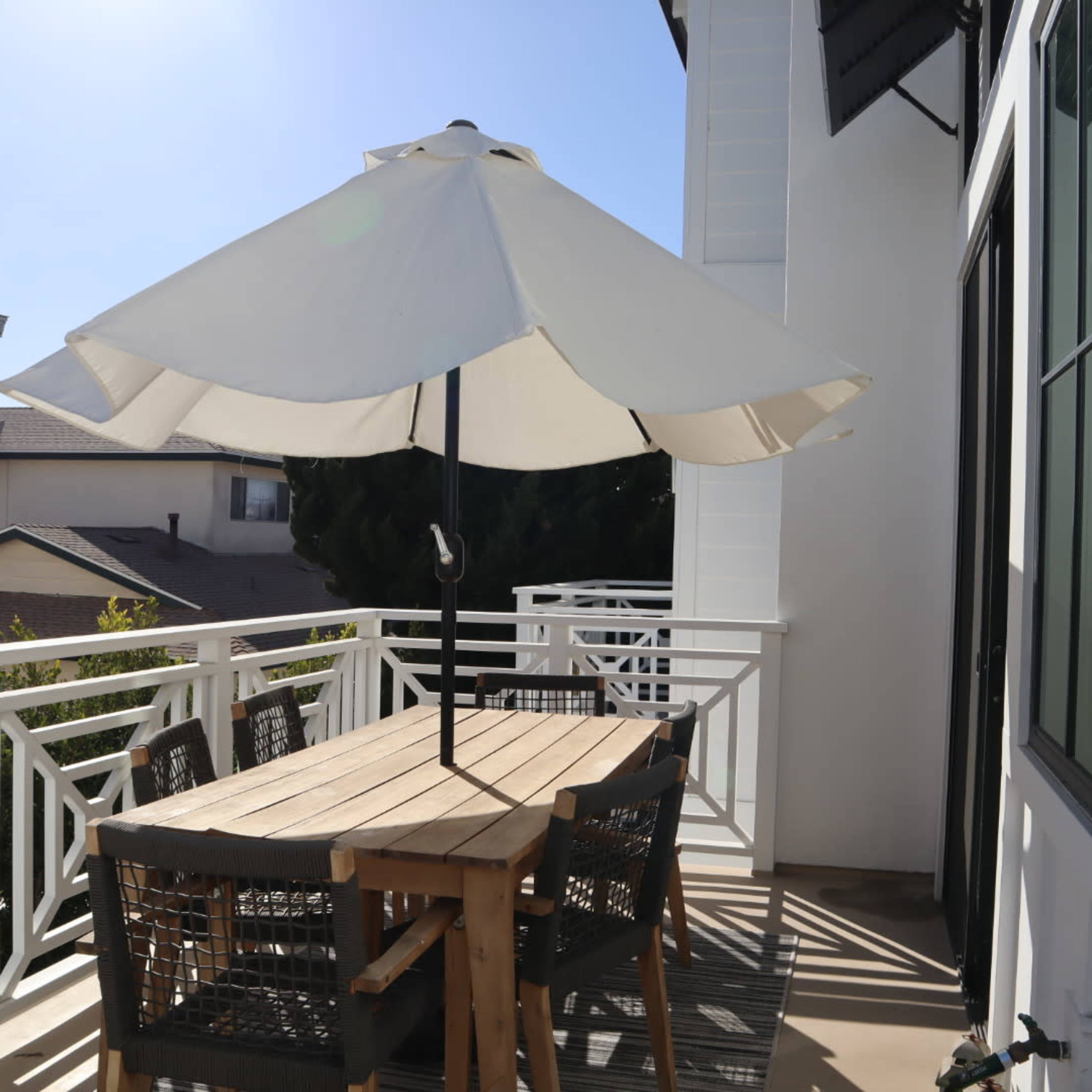 A patio area features a wooden table with wicker chairs and a large umbrella on a balcony overlooking nearby buildings.