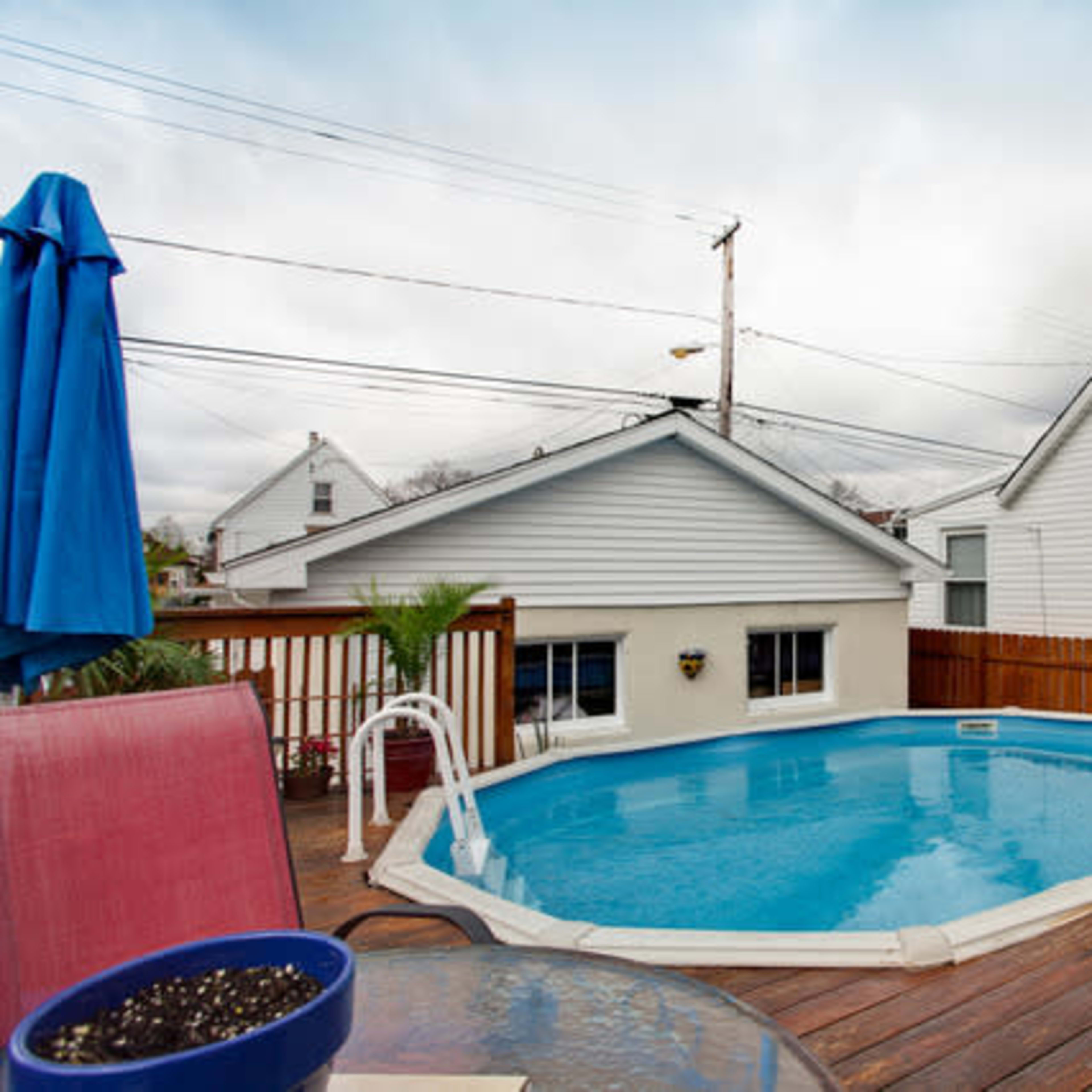 A small, blue above-ground pool is surrounded by a wooden deck and adjacent to a white house with a fence, under an overcast sky.