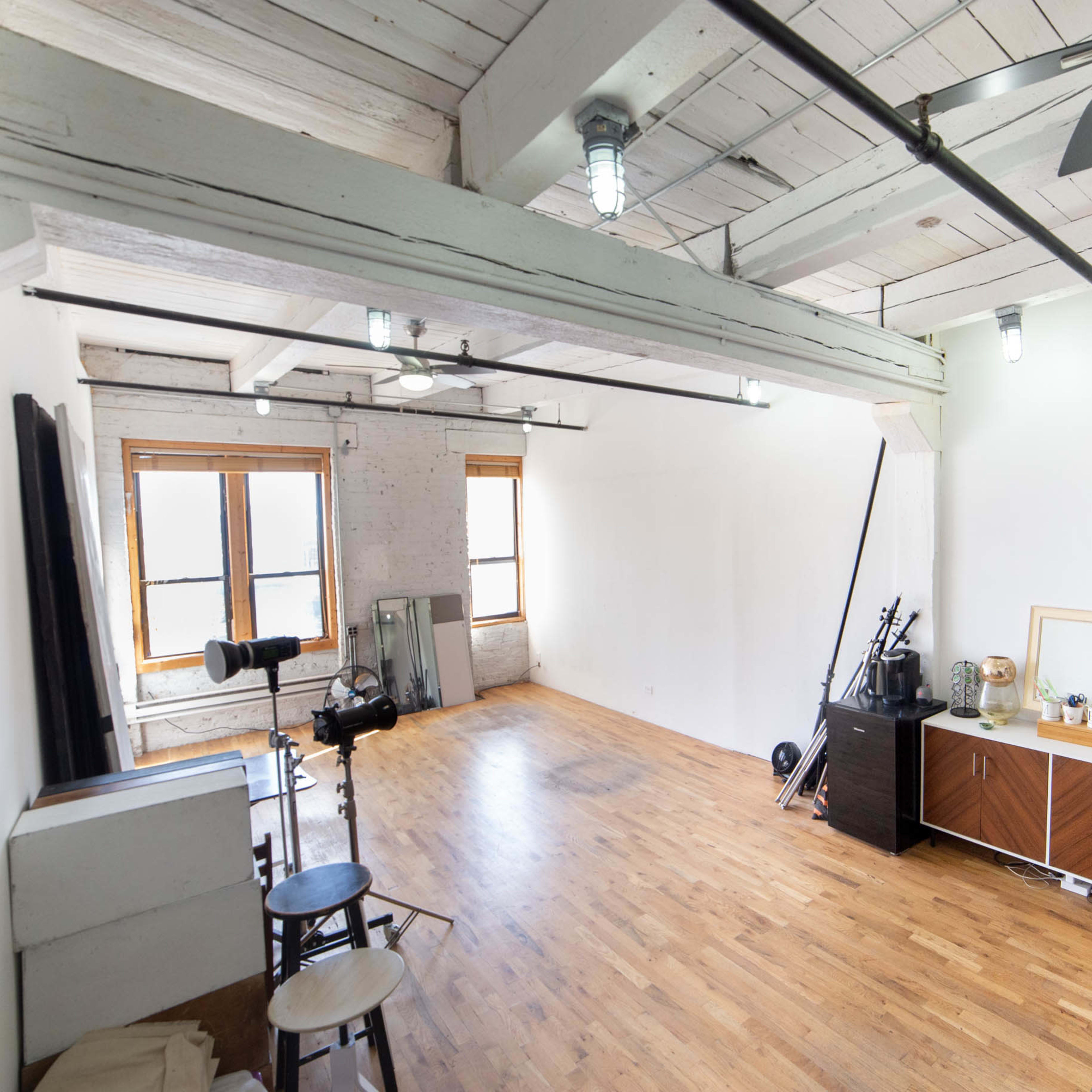 A spacious, empty room with wooden flooring, large windows, and minimal furnishings including a mirror, a camera setup, and storage furniture against the walls.