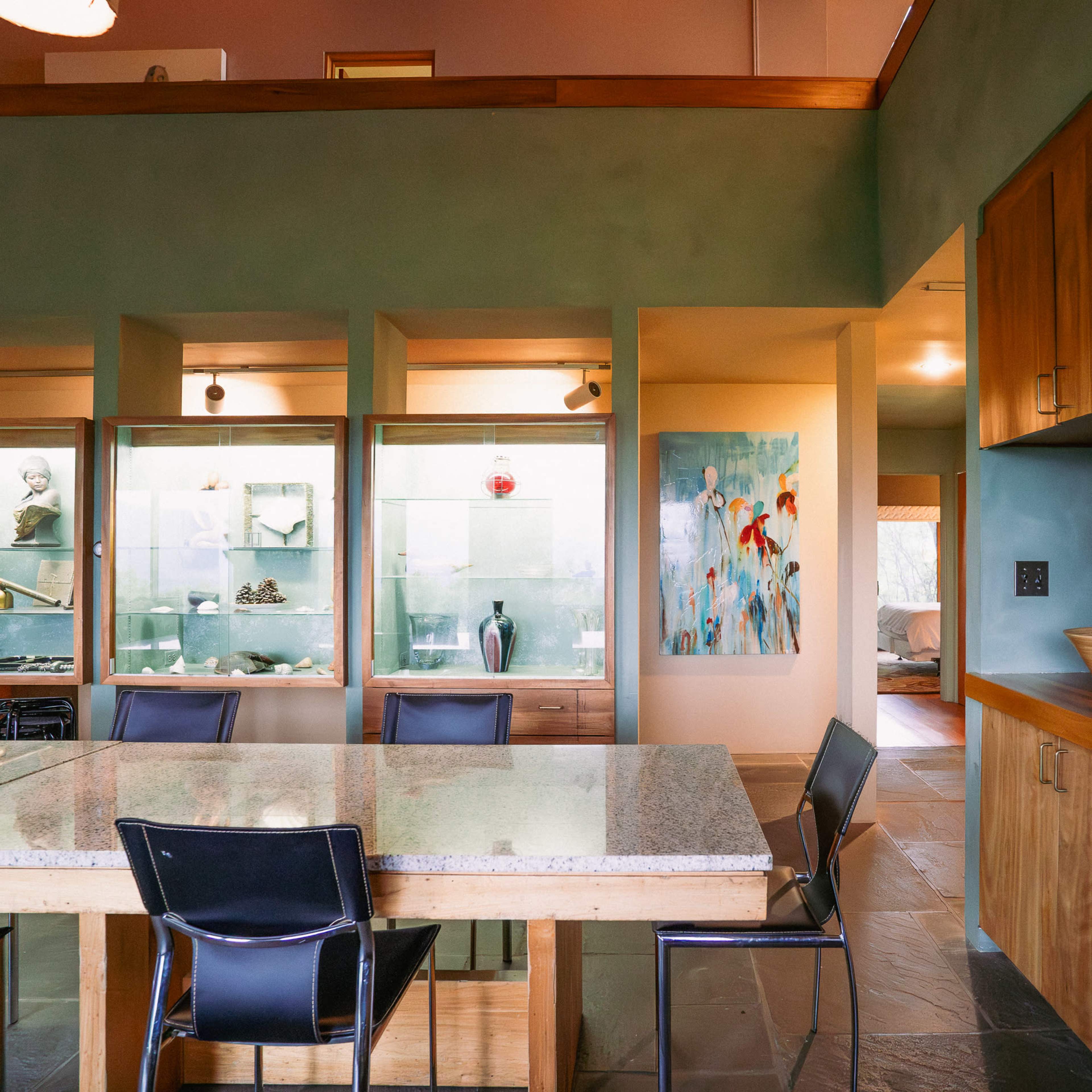 A modern dining area features a granite table surrounded by black chairs, with display cabinets showcasing decorative items and a colorful painting on the wall.