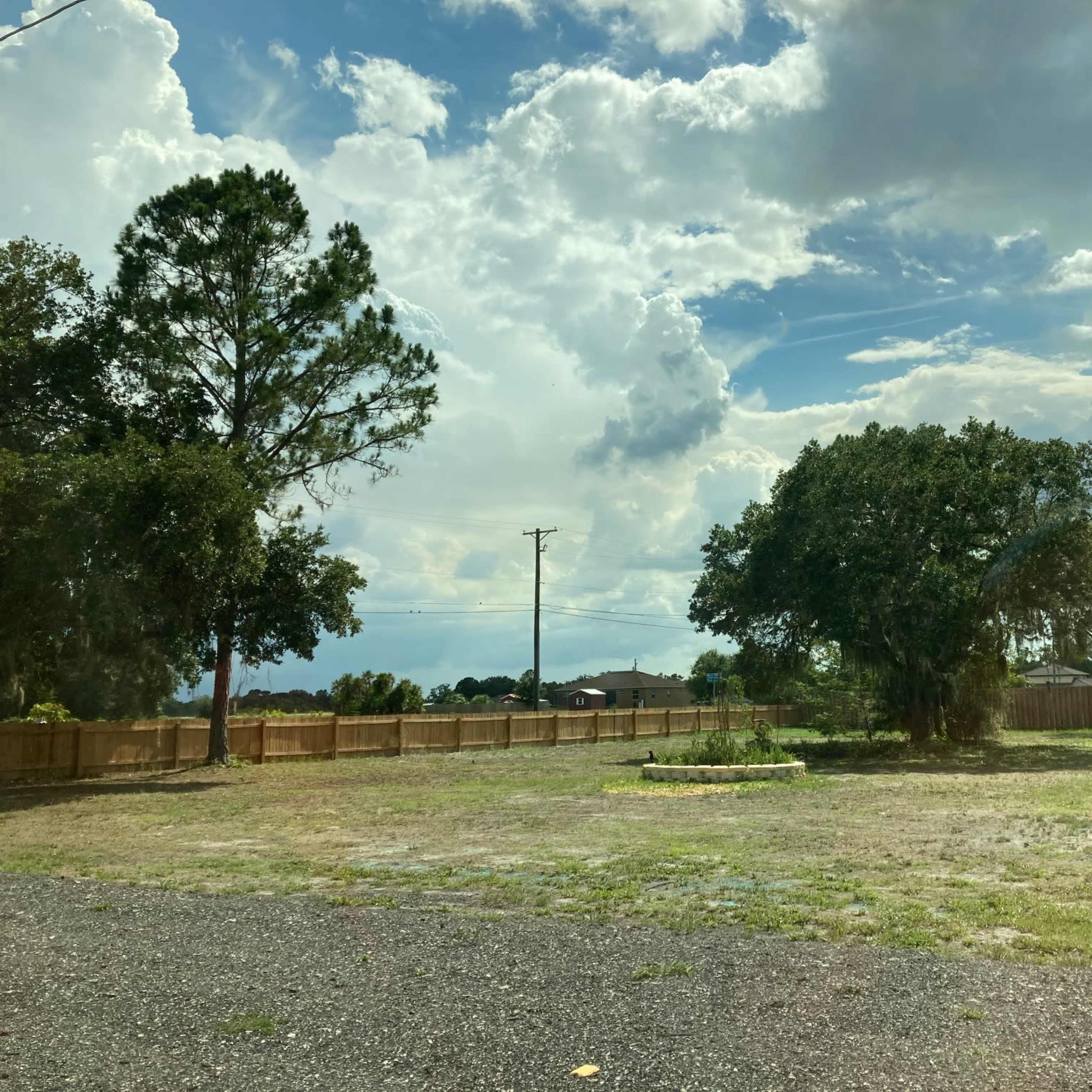 A grassy open area with a small circular garden feature, surrounded by a wooden fence and several trees under a cloudy sky.
