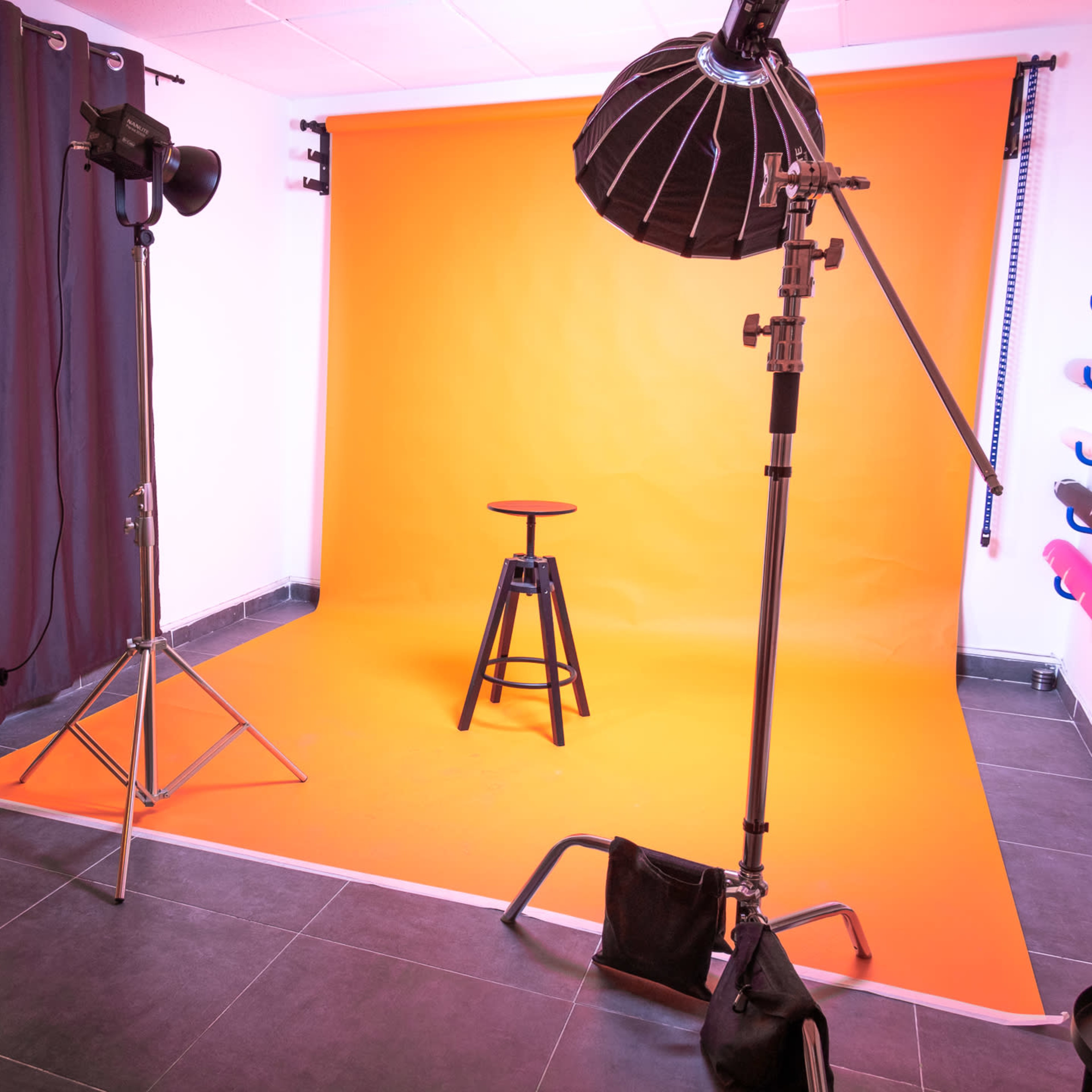 A photography studio featuring a wooden stool in front of an orange backdrop, with lighting equipment and a reflector positioned nearby.