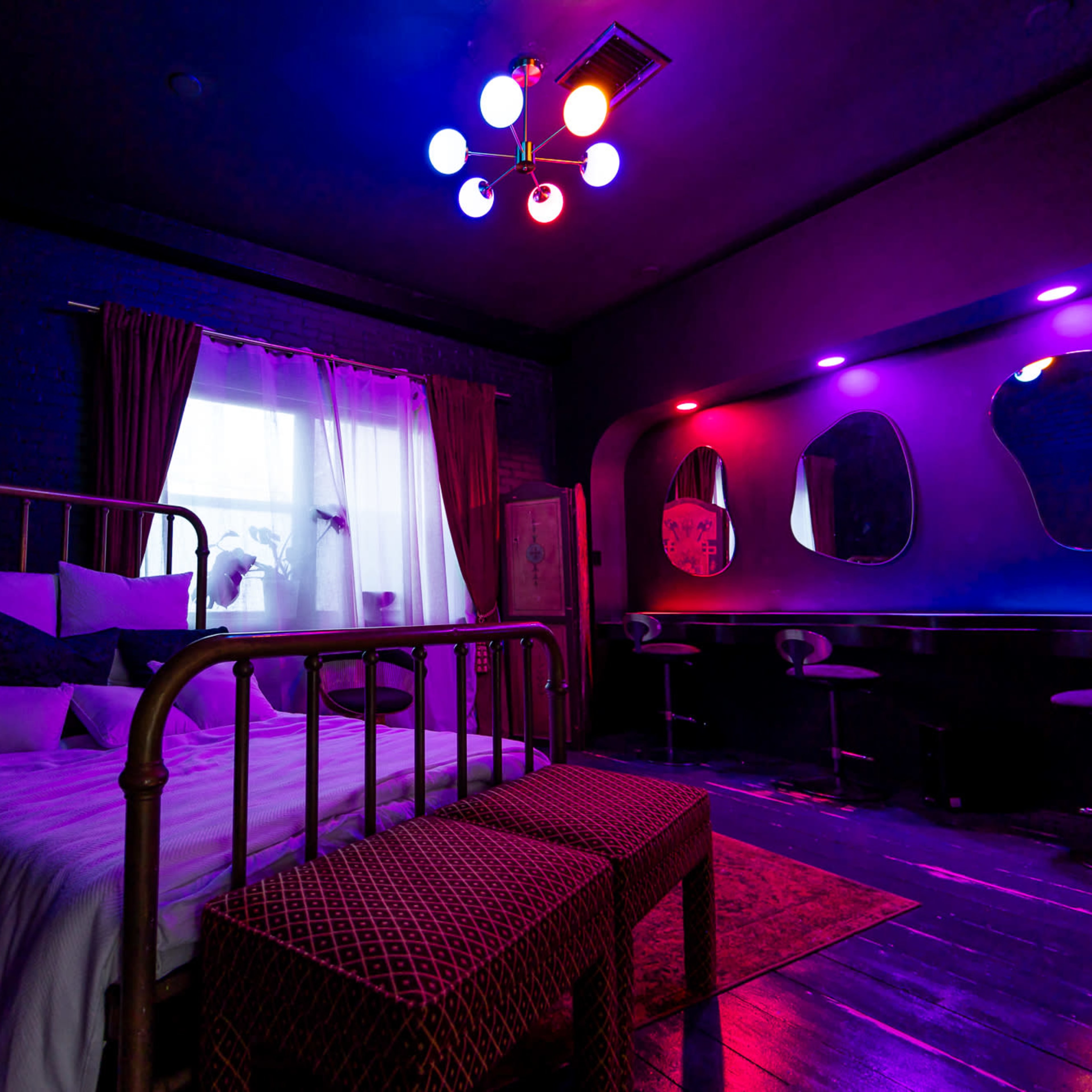 A bedroom features a metallic bed with a white bedspread, vibrant lighting, and a modern seating area with two mirrors.