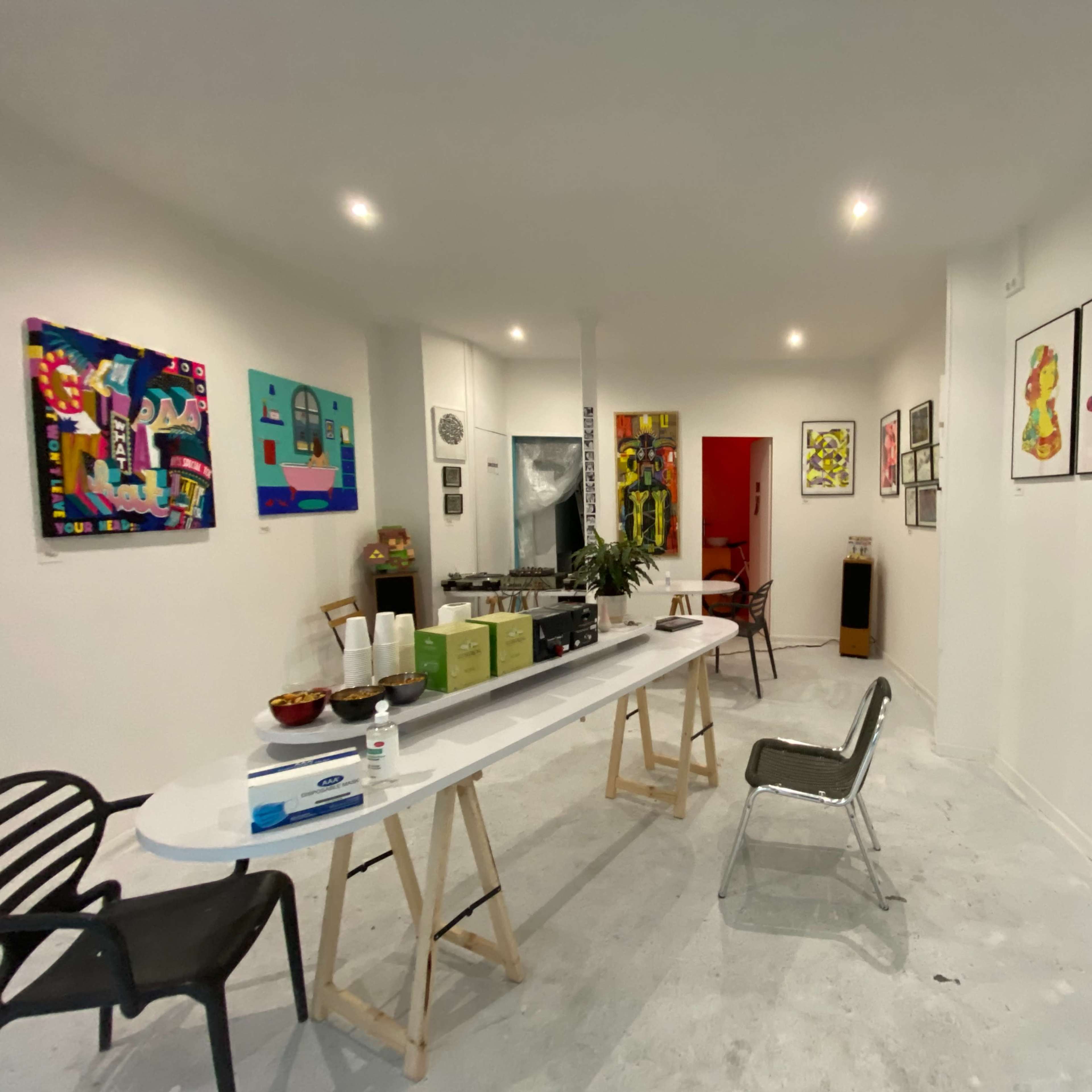 An art gallery featuring colorful paintings on the walls, with a table set up in the center displaying refreshments.