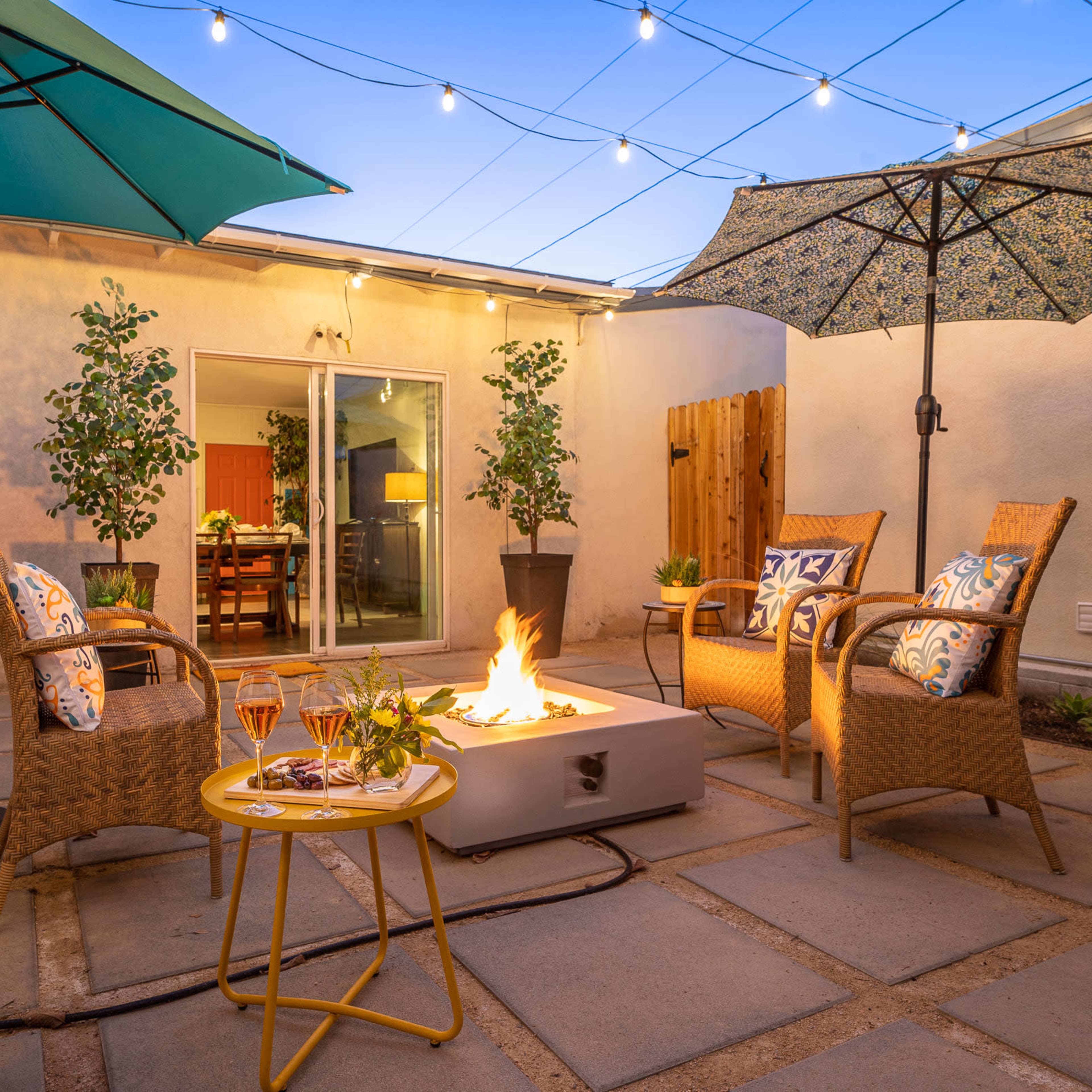 A cozy outdoor seating area with four chairs around a fire pit, surrounded by umbrellas and small plants, illuminated by string lights overhead.