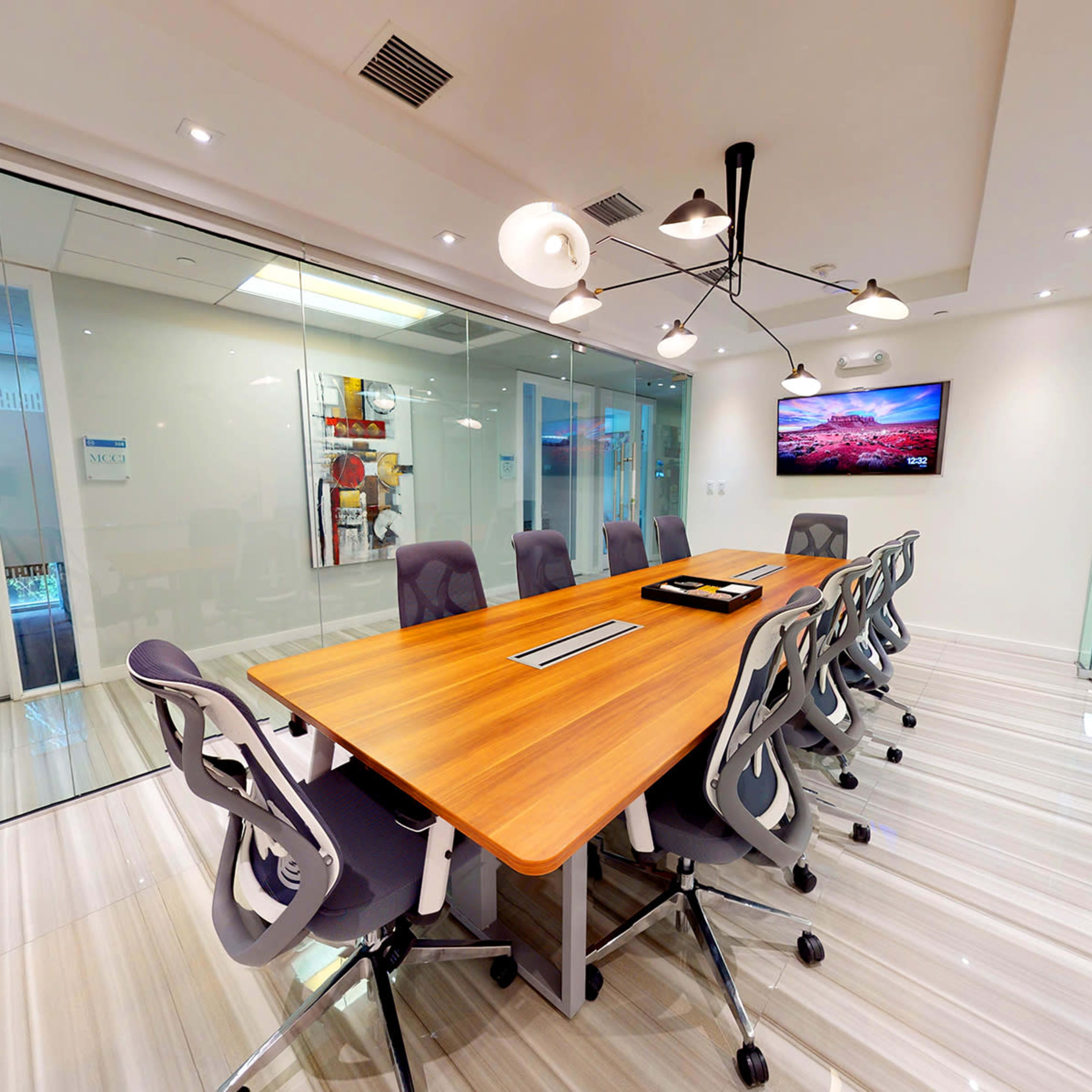 A modern conference room features a long wooden table surrounded by ergonomic chairs, with glass walls and a large artwork on display.