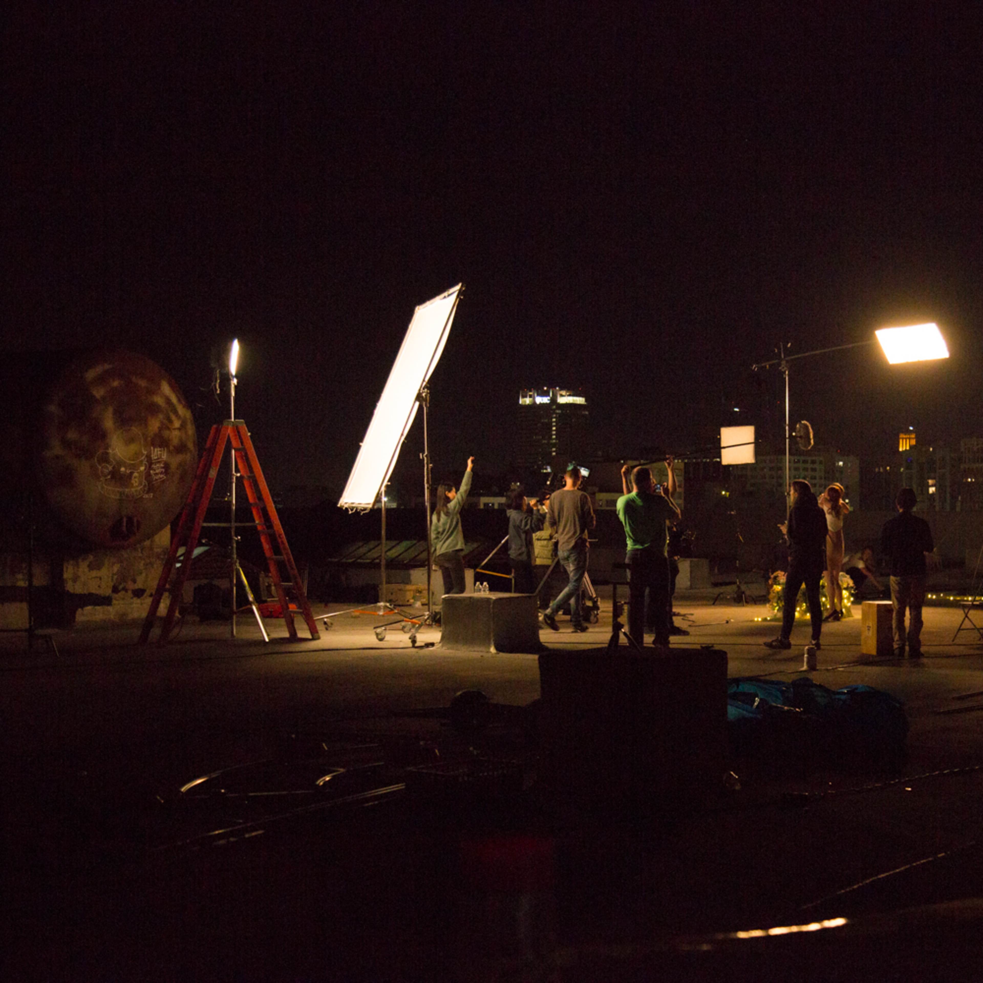 A film crew sets up lighting equipment on a rooftop at night, with city lights visible in the background.