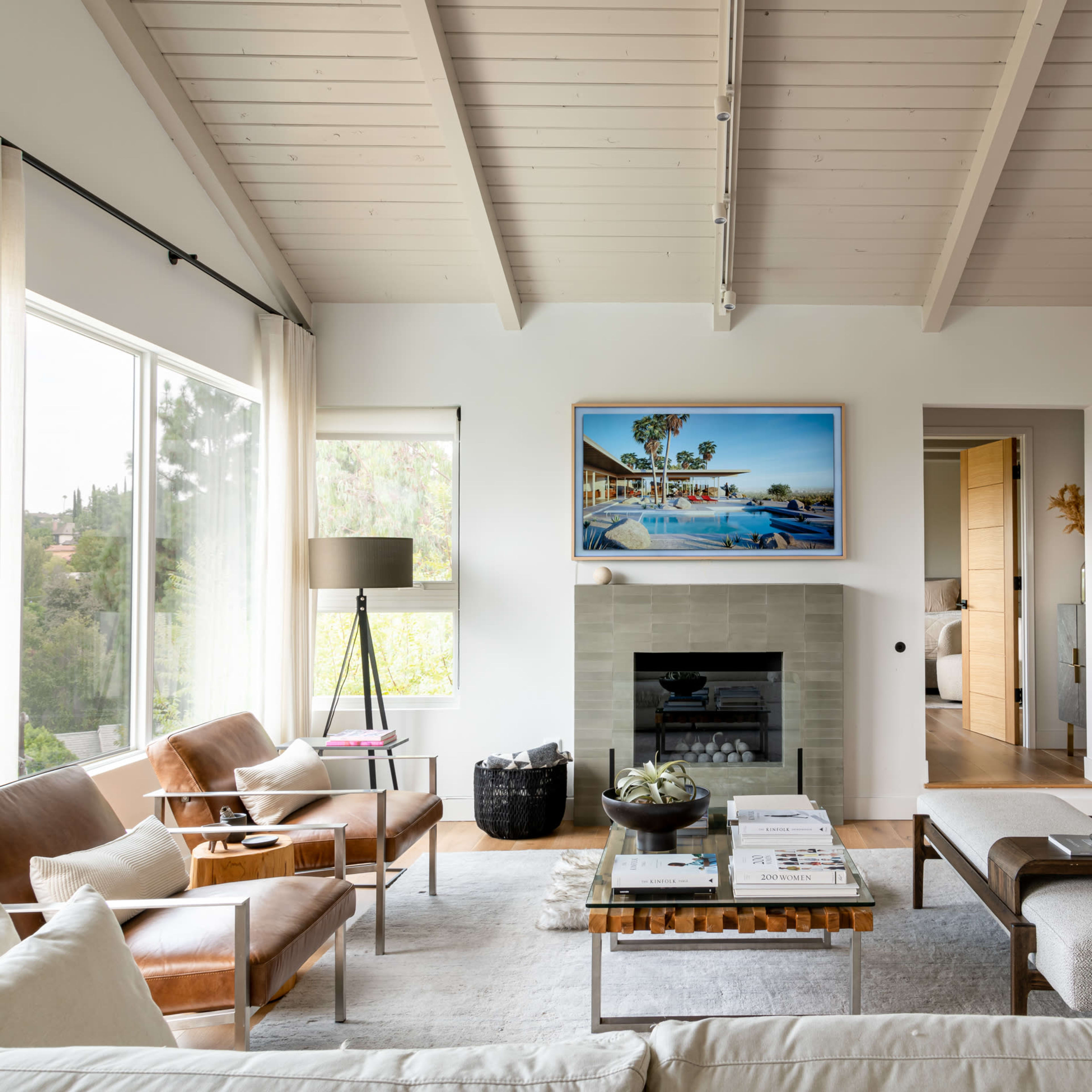 A modern living room with a fireplace, large windows, and a couch, featuring neutral tones and wooden ceiling beams.