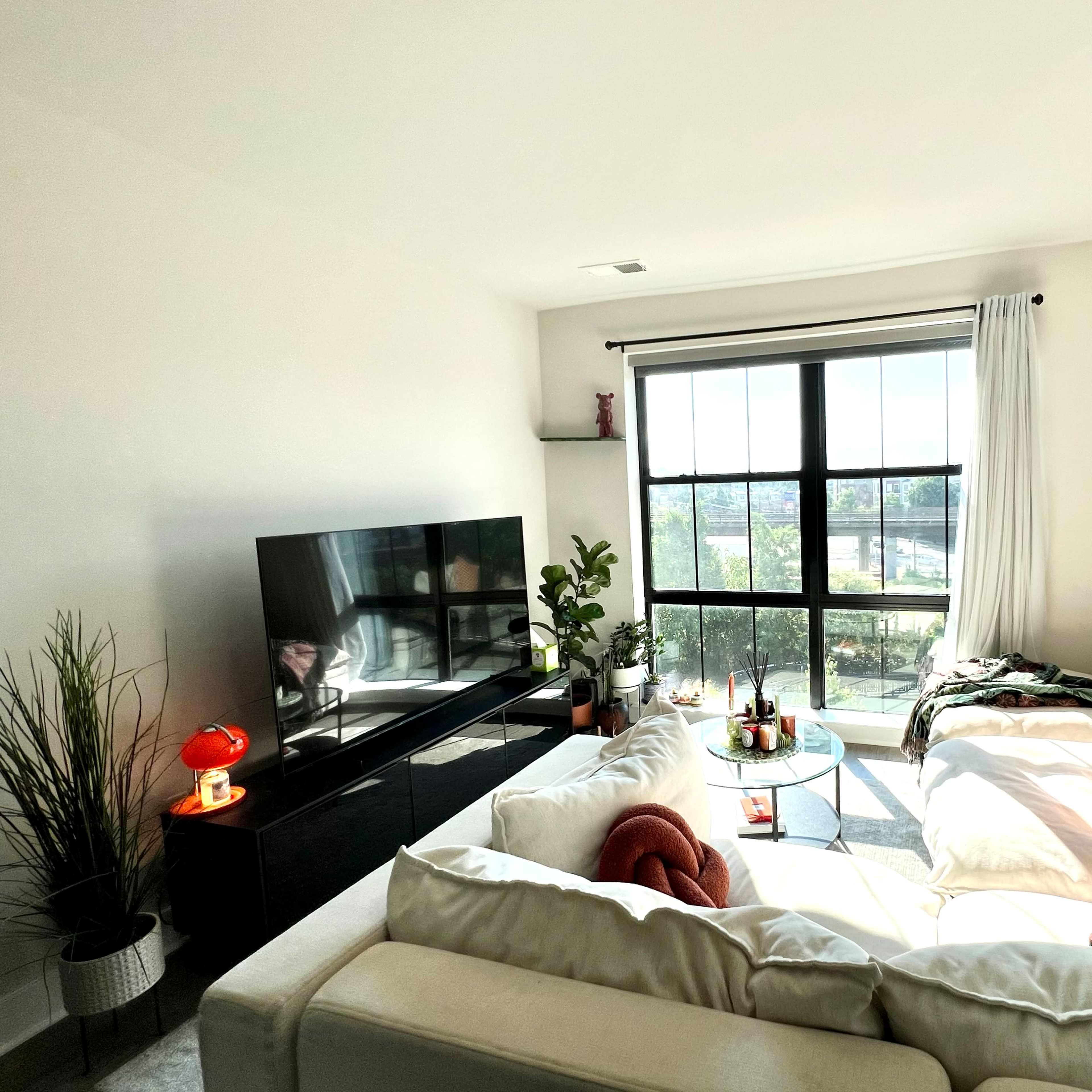 A bright living room features a sectional sofa, a coffee table, a large television, and a window with a view.