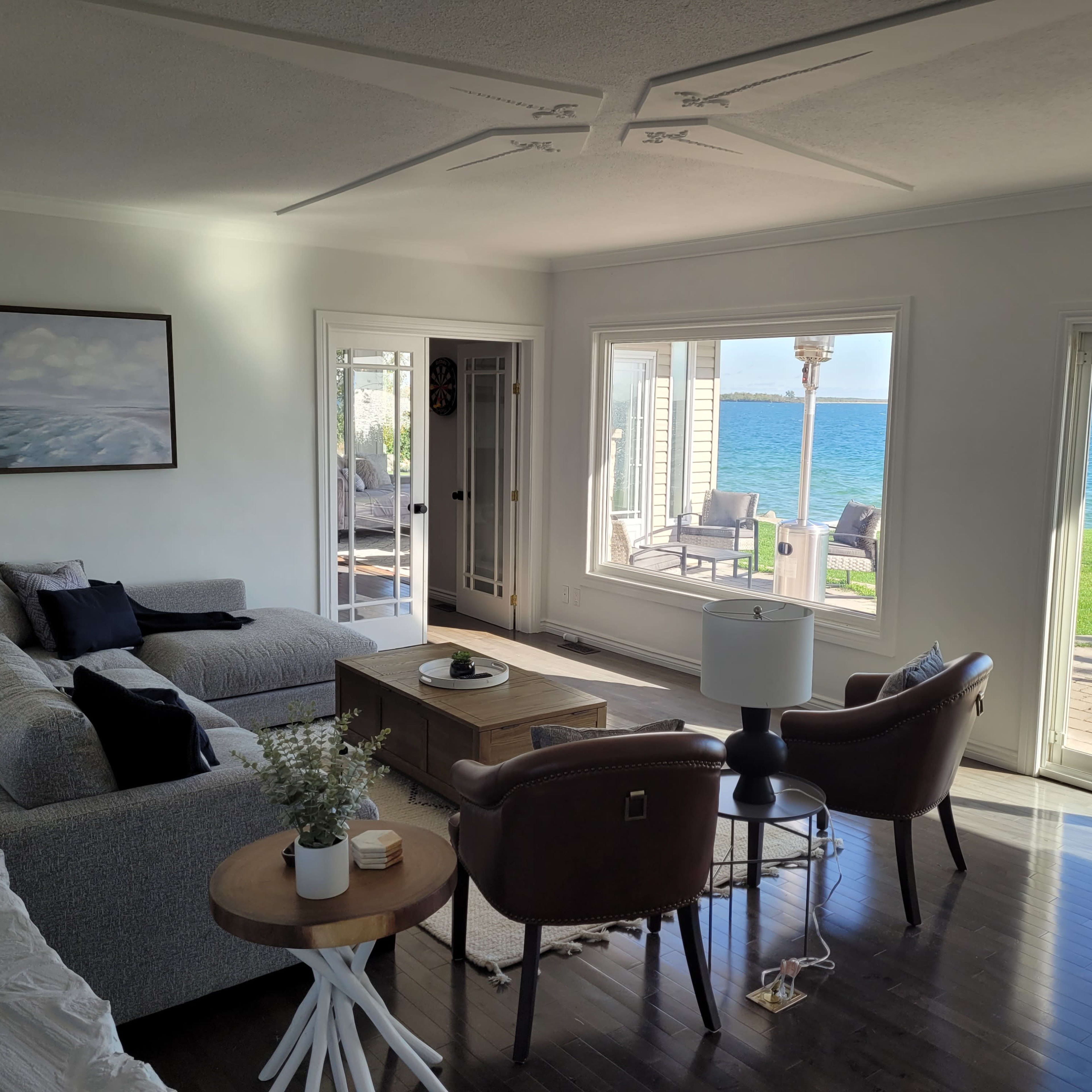 A modern living room features a gray sofa, two brown armchairs, and large windows offering a view of the water.