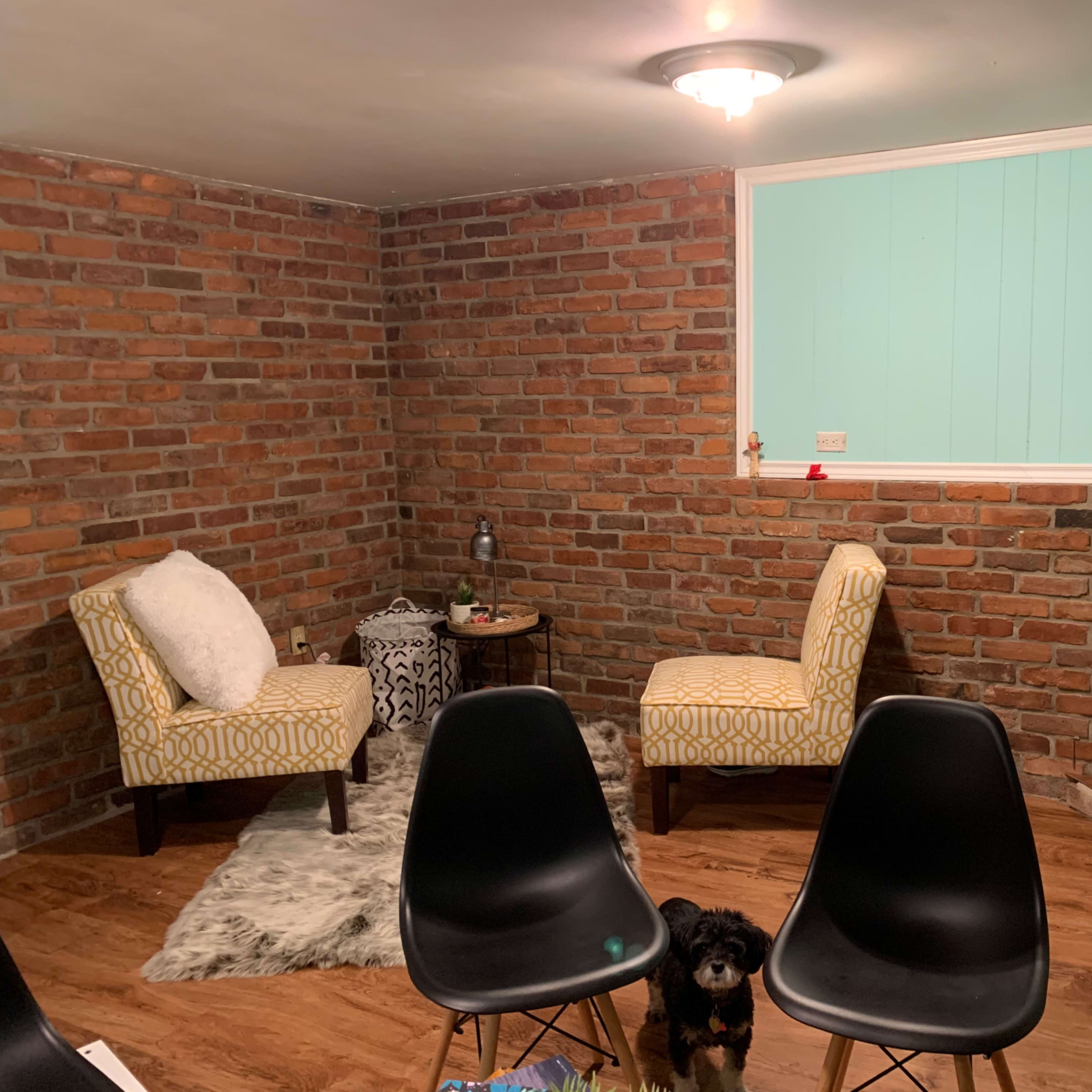 A cozy room with exposed brick walls, two patterned chairs, and a dog on a wooden floor, accompanied by a fireplace and a small table.