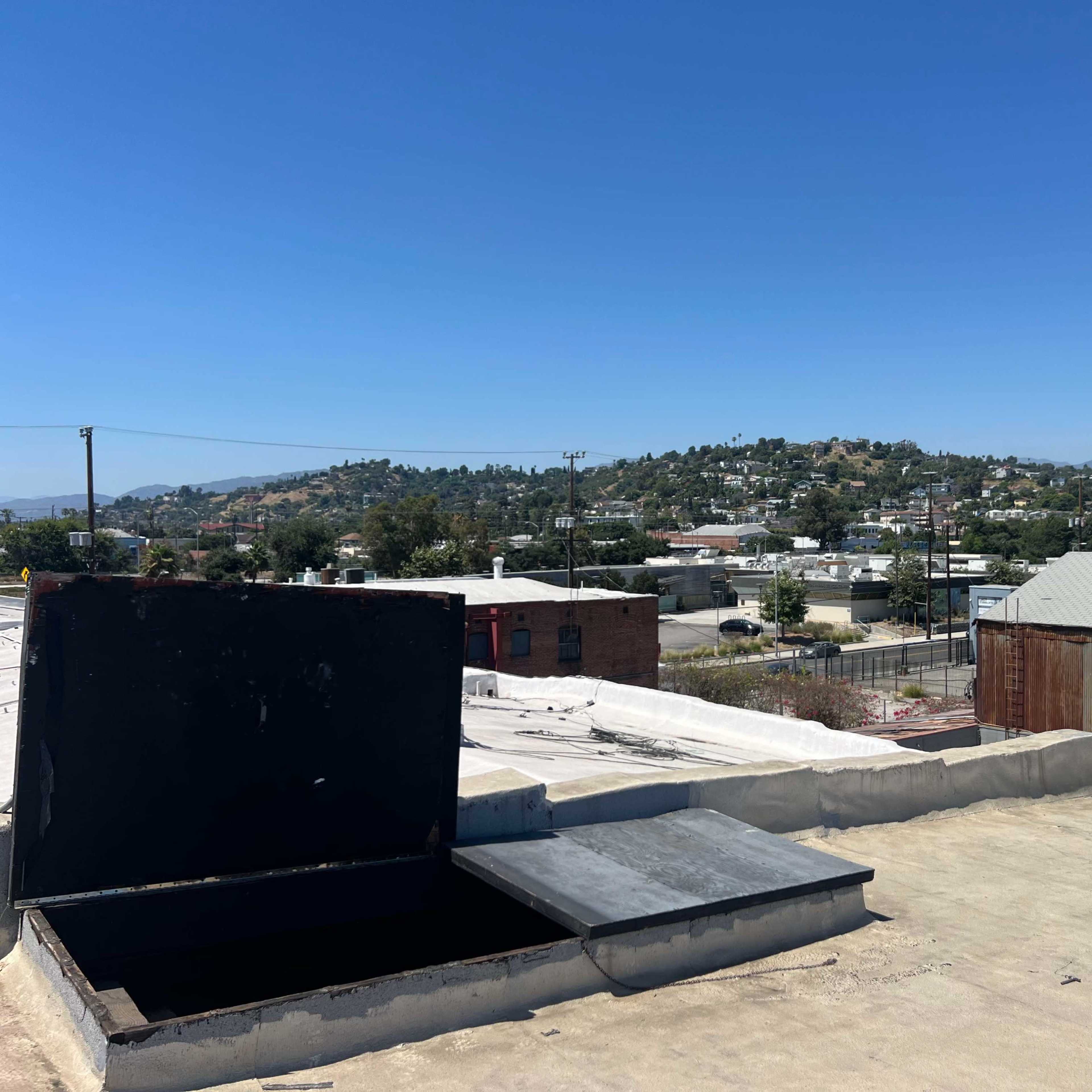 A rooftop with an open hatch and a view of a hillside and buildings in the background under a clear blue sky.
