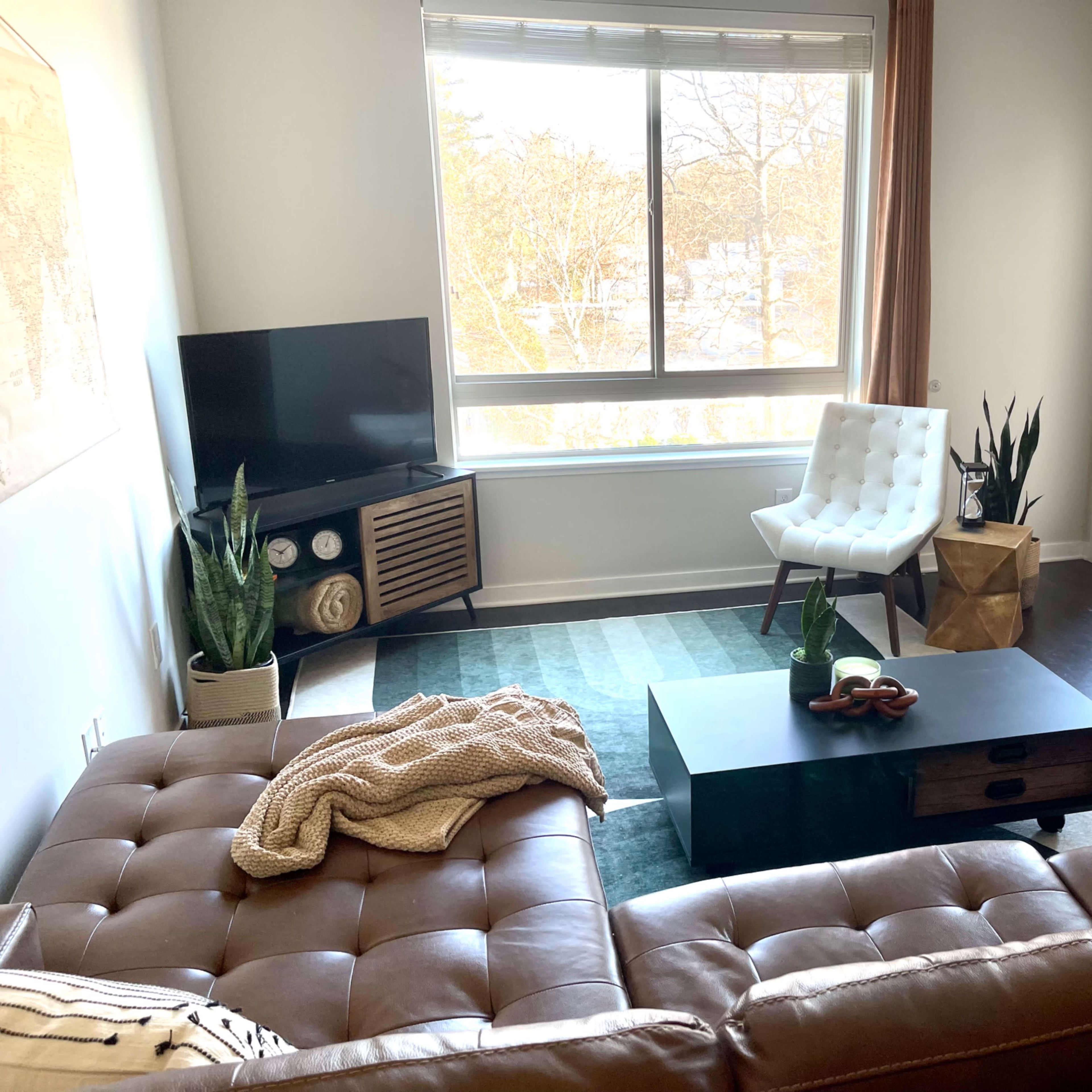 A modern living room with a brown leather sofa, a coffee table, a white chair, a television, and a view of a balcony through large windows.