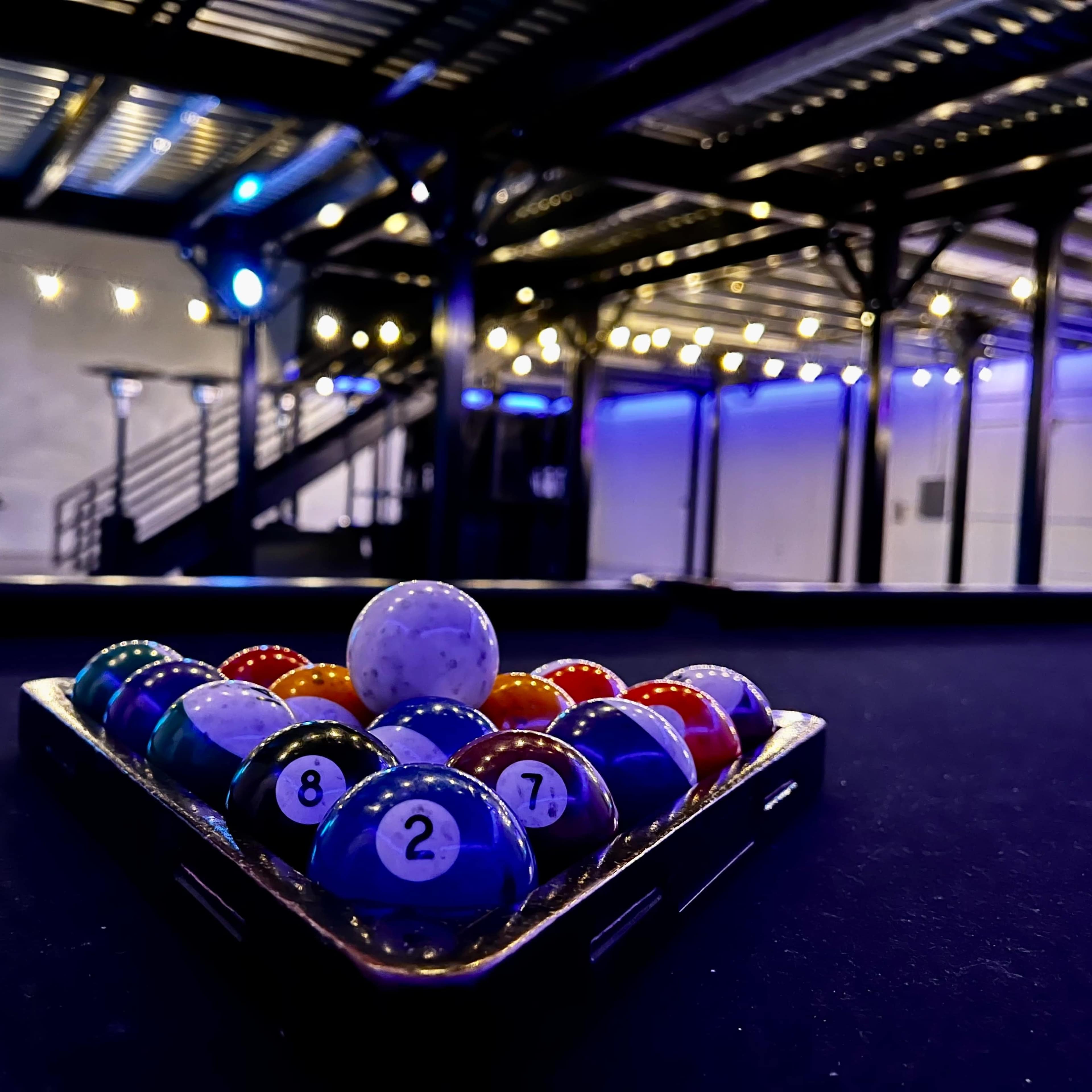 A triangular rack of colorful billiard balls is positioned on a pool table in a dimly lit room with string lights overhead.
