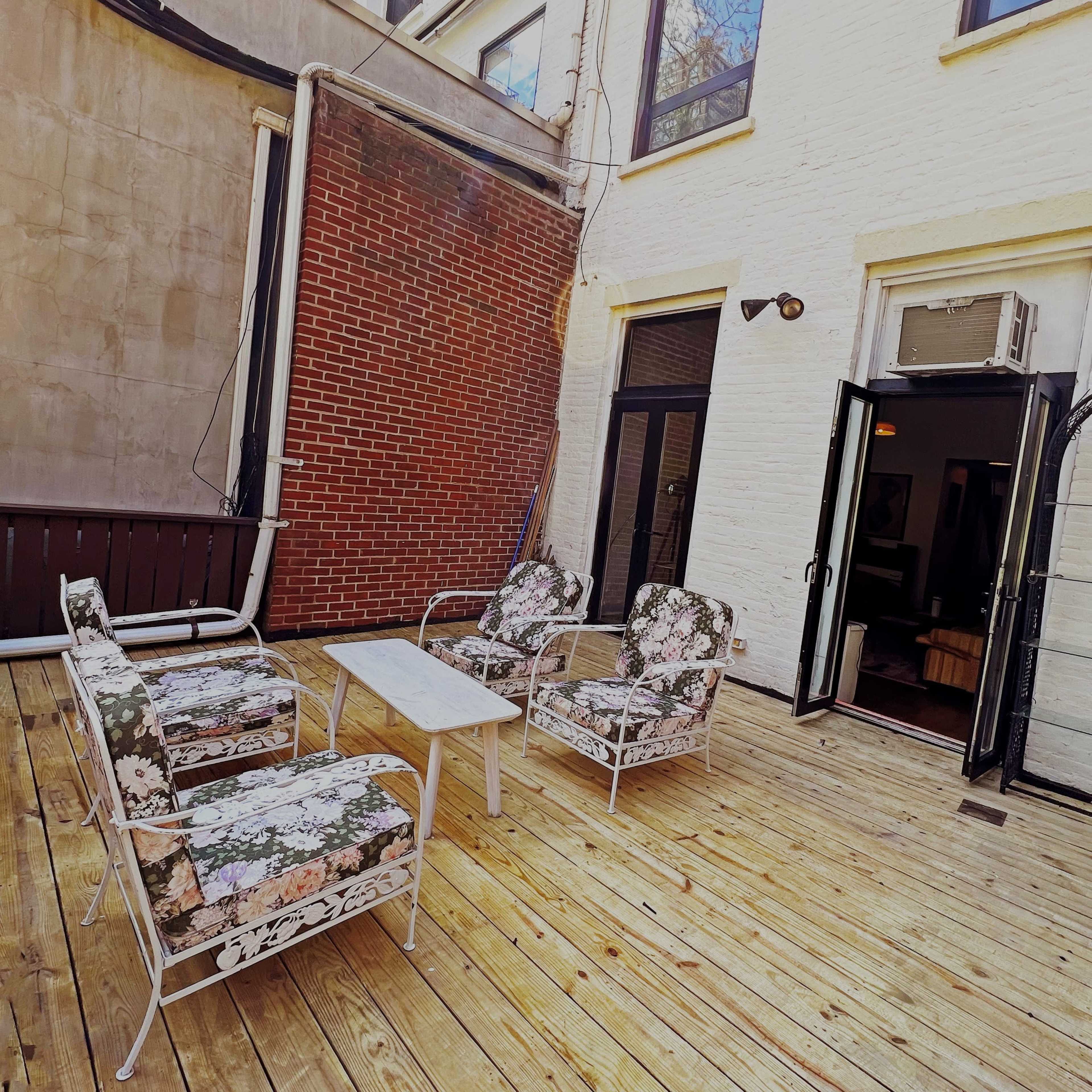 A wooden deck features four floral-patterned chairs and a small table, adjacent to an open doorway leading inside.