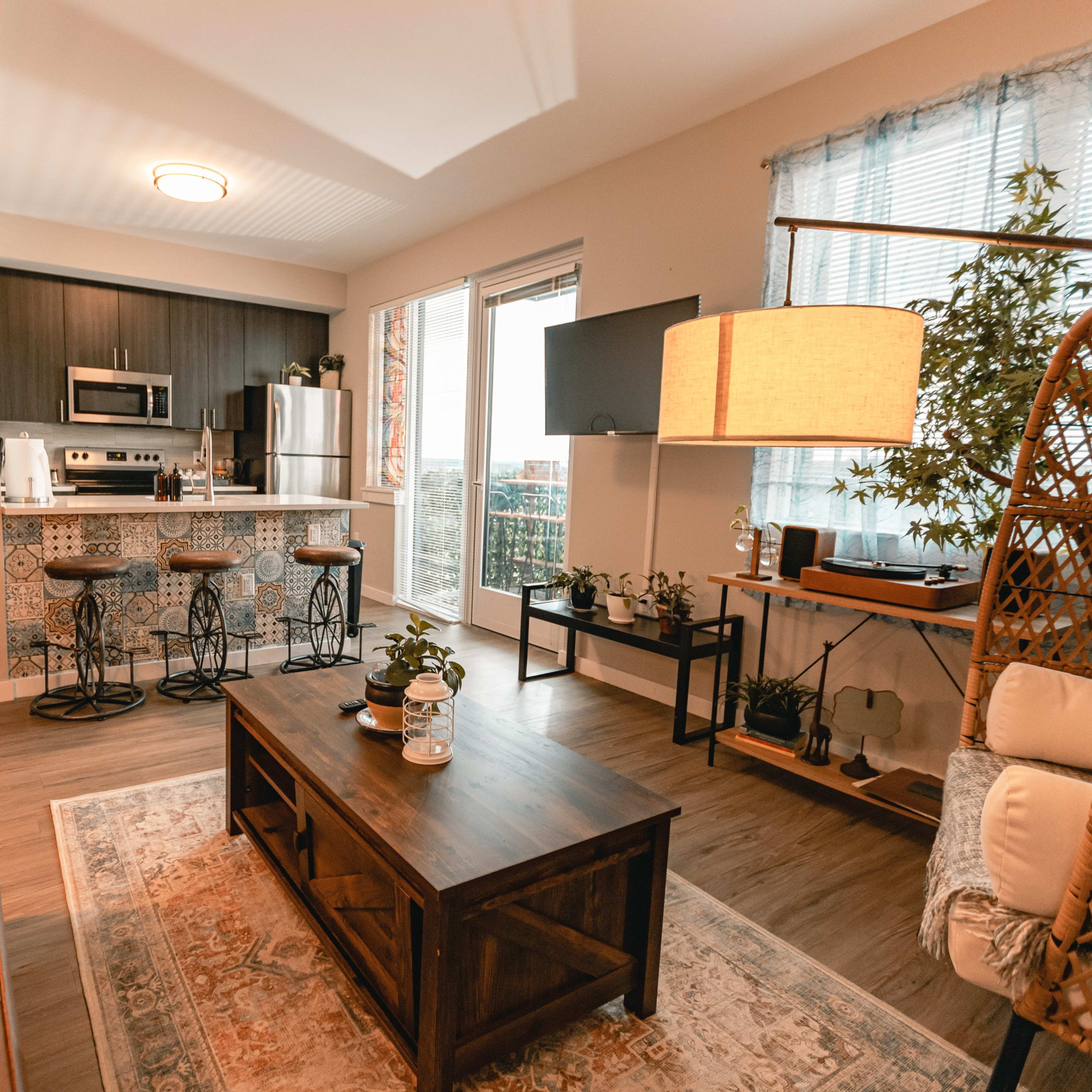 A modern living room with a brown leather couch, a wooden coffee table, a kitchen in the background featuring bar stools, and large windows with natural light.