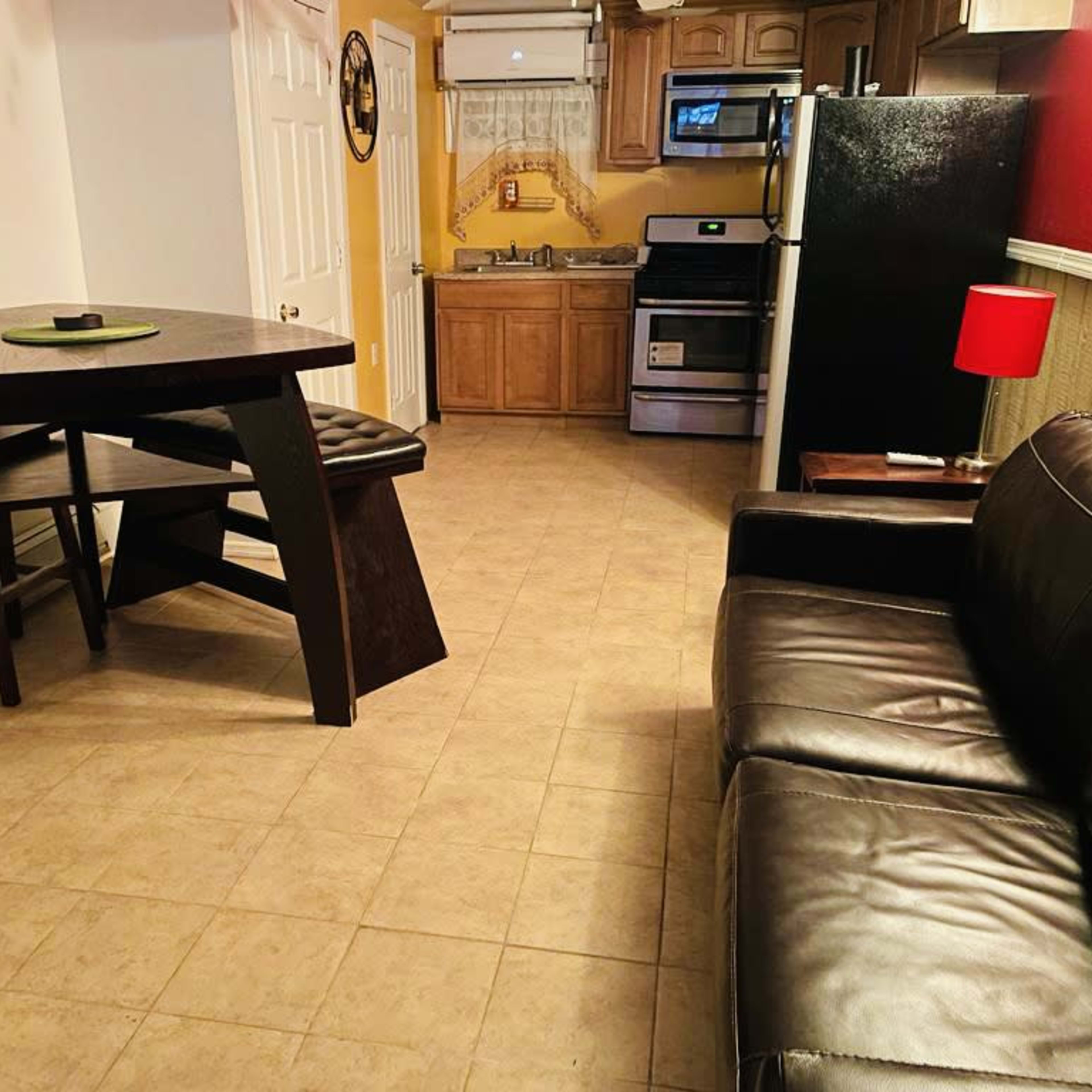 A small kitchen and living area featuring a black table with stools, a leather couch, and kitchen appliances, including a refrigerator and stove.