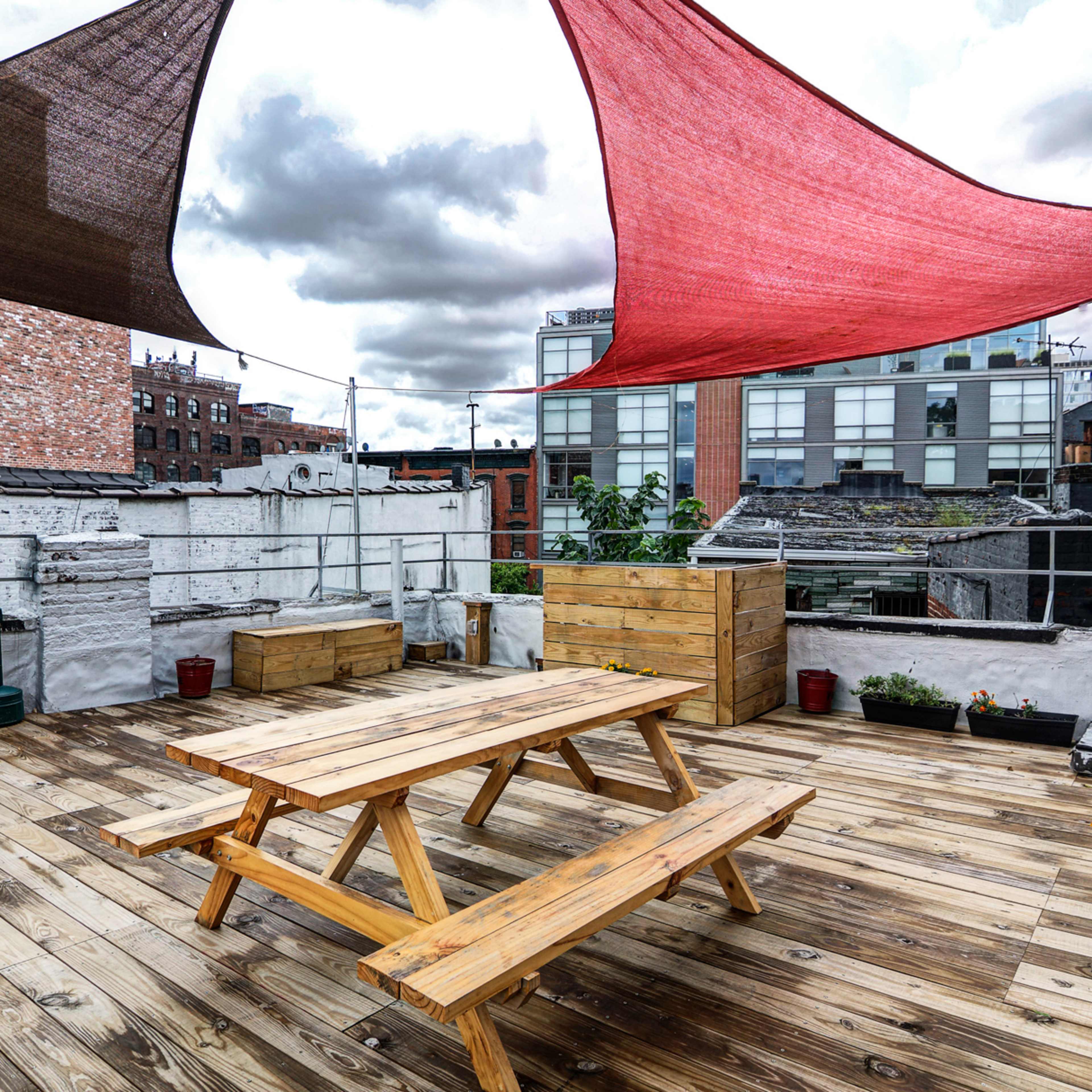 A rooftop terrace features a wooden table and benches beneath colorful shade sails, with planters and a grill set against a backdrop of urban buildings.