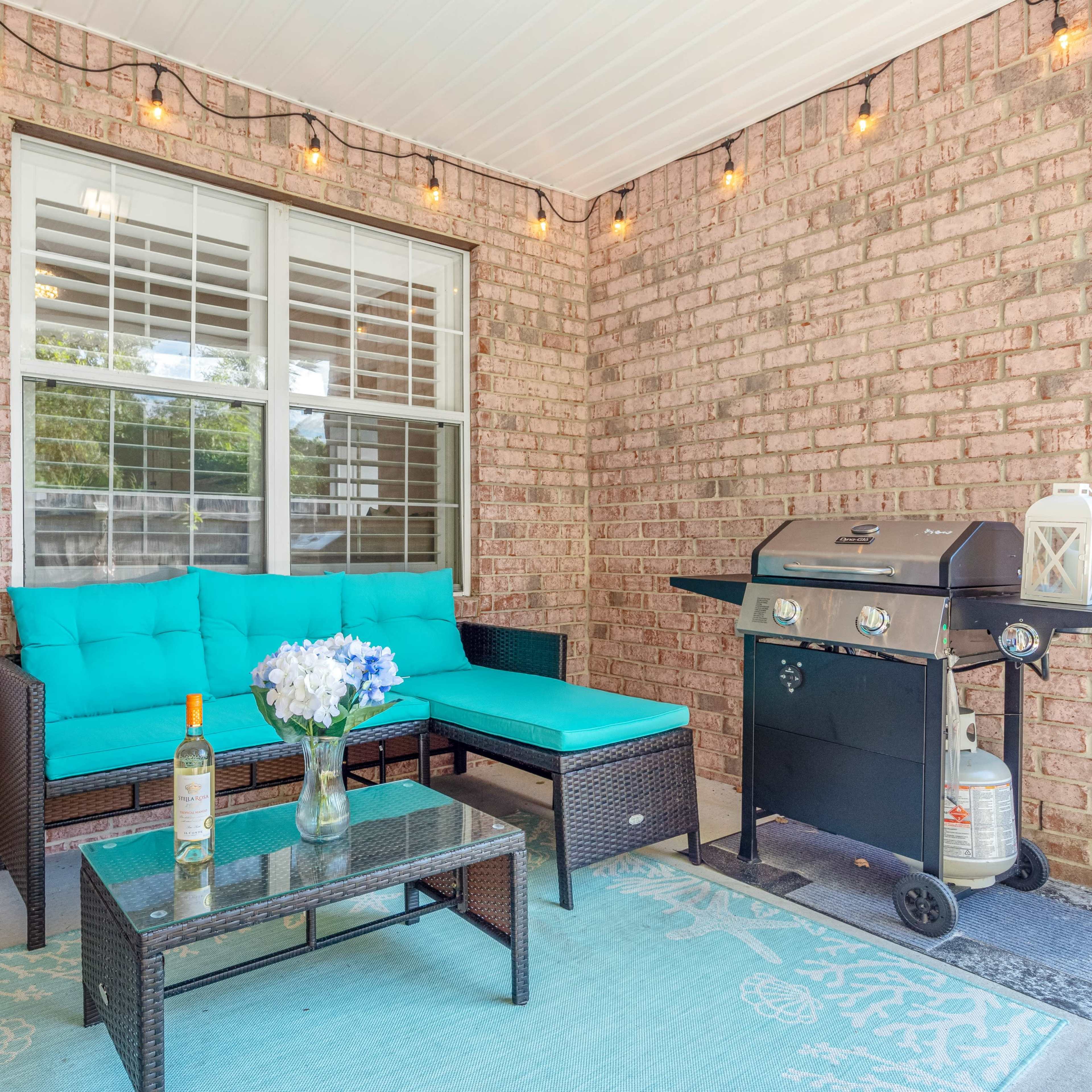 A covered patio with a turquoise seating area, a glass coffee table, and a charcoal grill beside it.