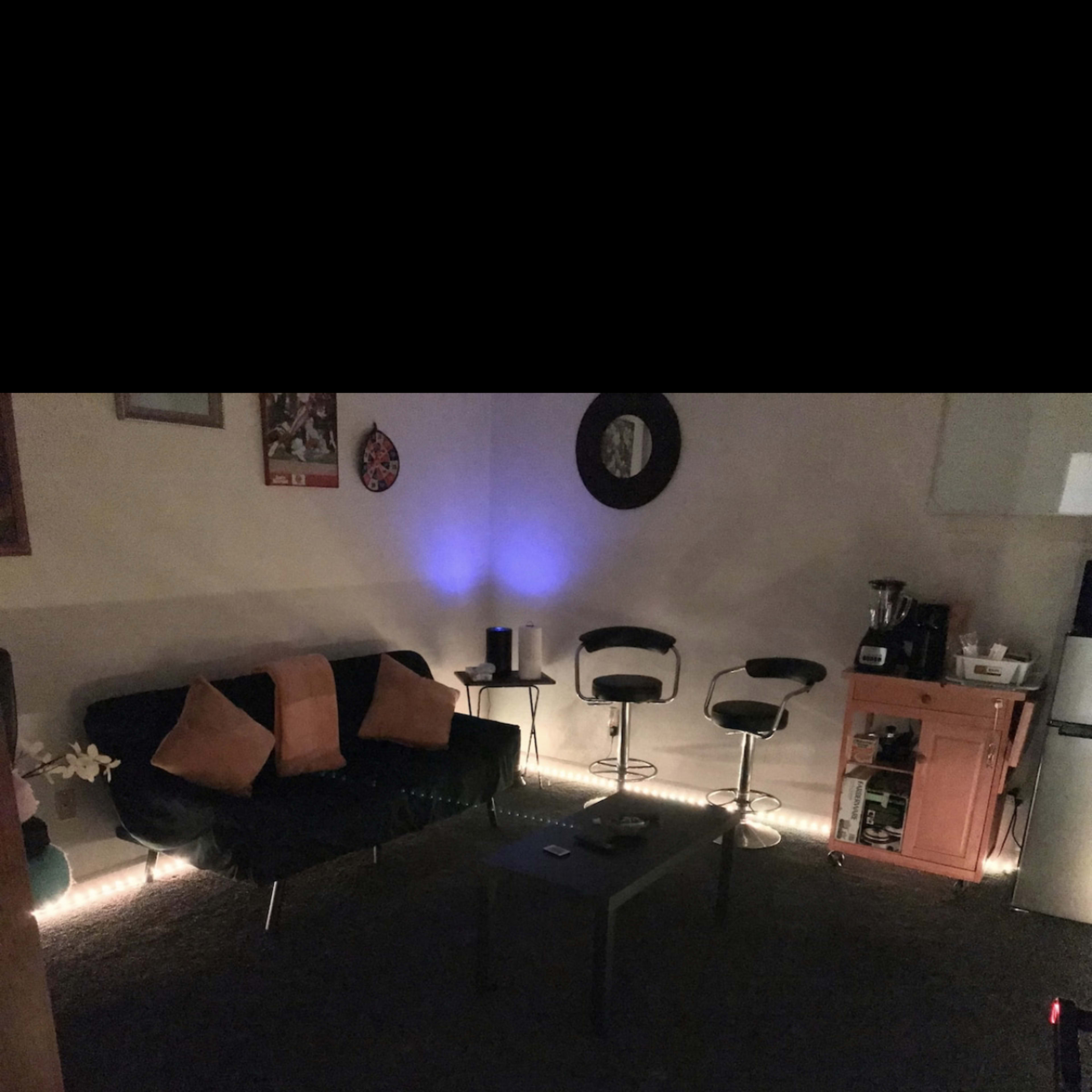 A dark living area with a black couch, four pillows, a coffee table, two bar stools, and a kitchenette visible in the background.