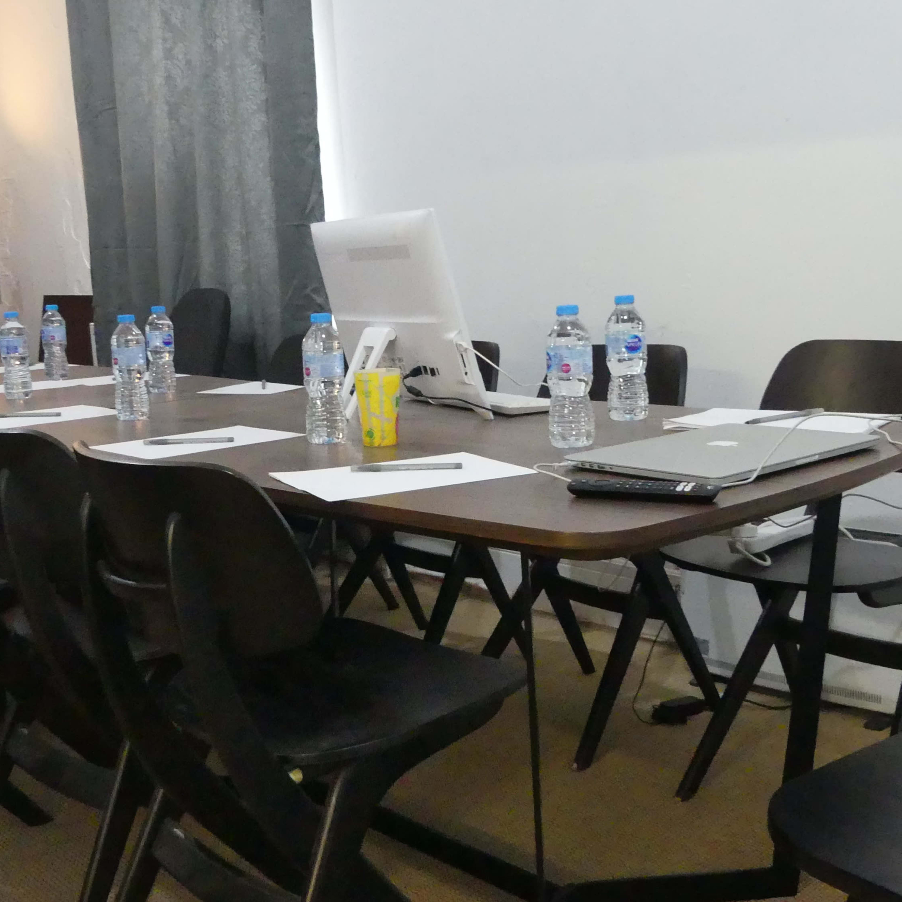 A rectangular table is set up in a room with several chairs around it, featuring water bottles, papers, and a computer monitor.