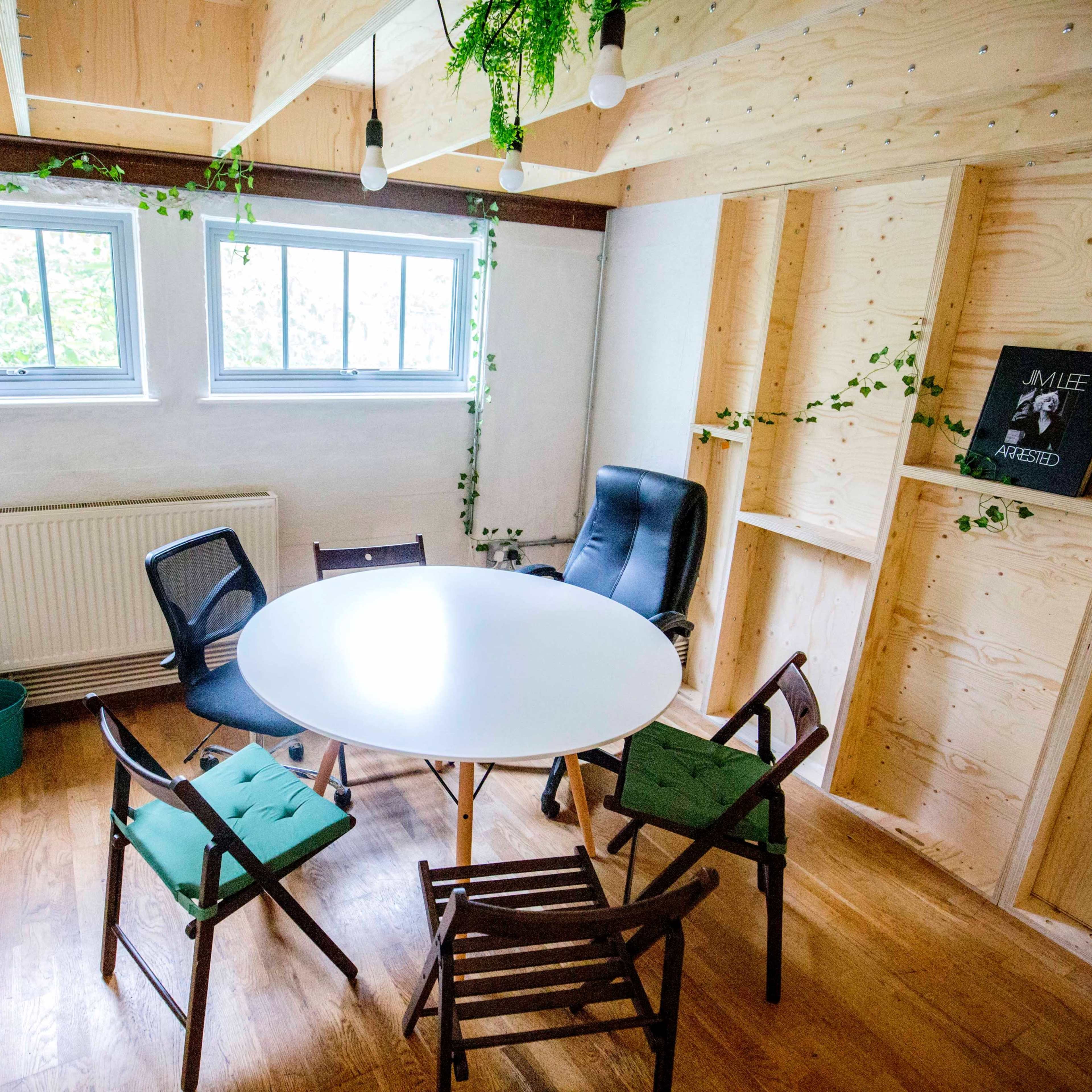 A small, bright room features a round white table surrounded by wooden chairs, with shelves and greenery adorning the walls.