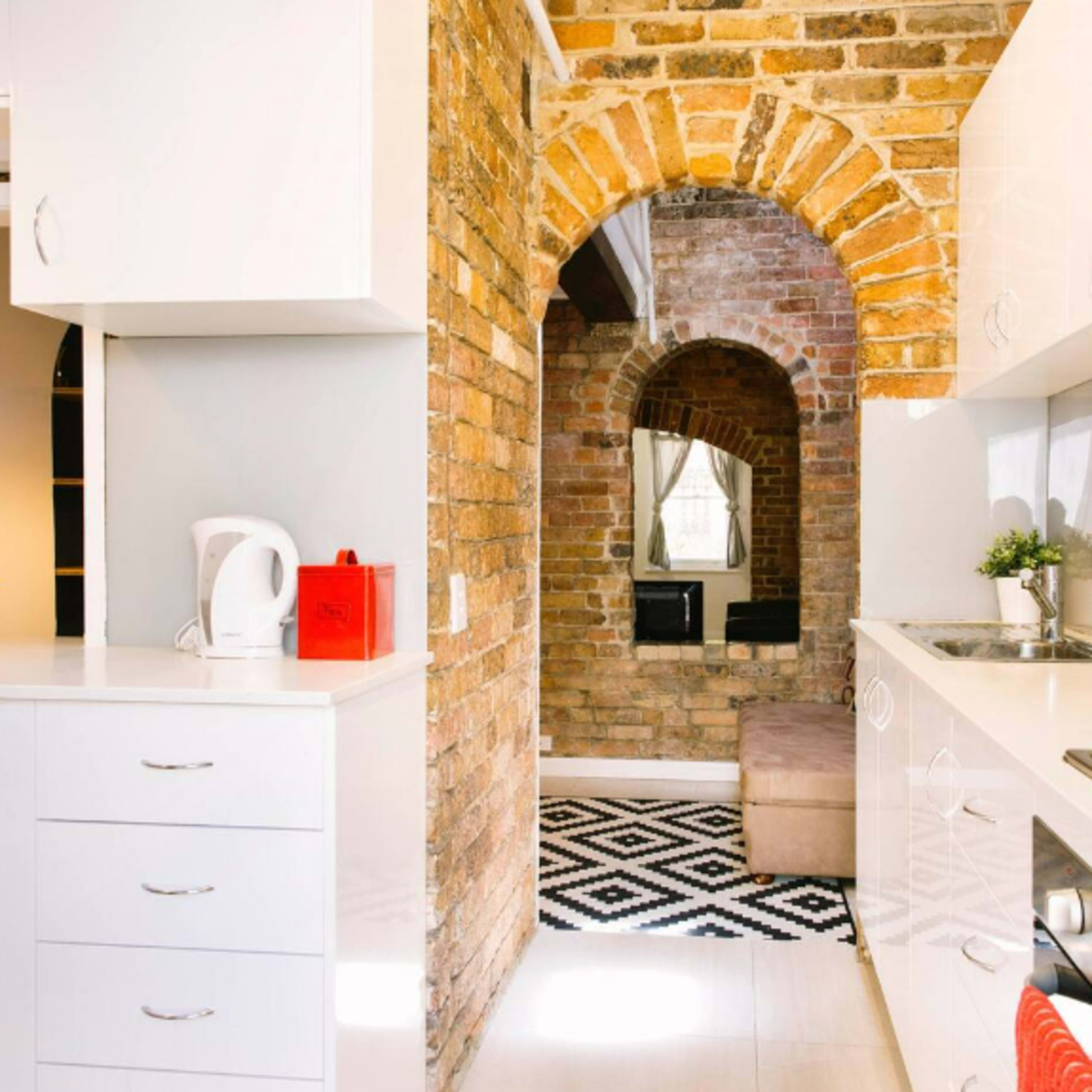 A modern kitchen with white cabinetry and a brick archway leading to a cozy living area.