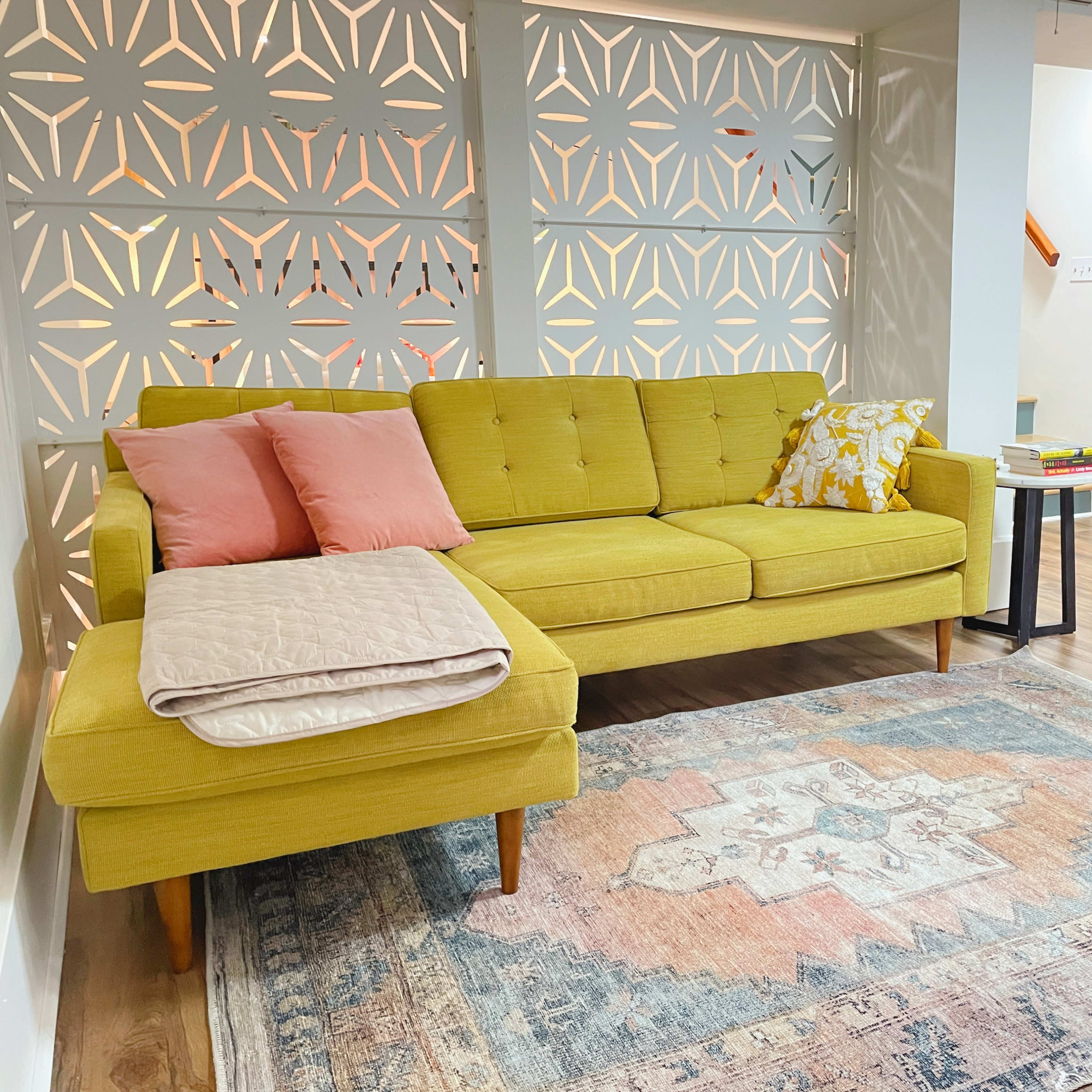 A mid-century modern yellow sectional sofa with pink and patterned cushions, placed on a patterned area rug in a spacious room with decorative partition screens and a glimpse of a bedroom in the background.