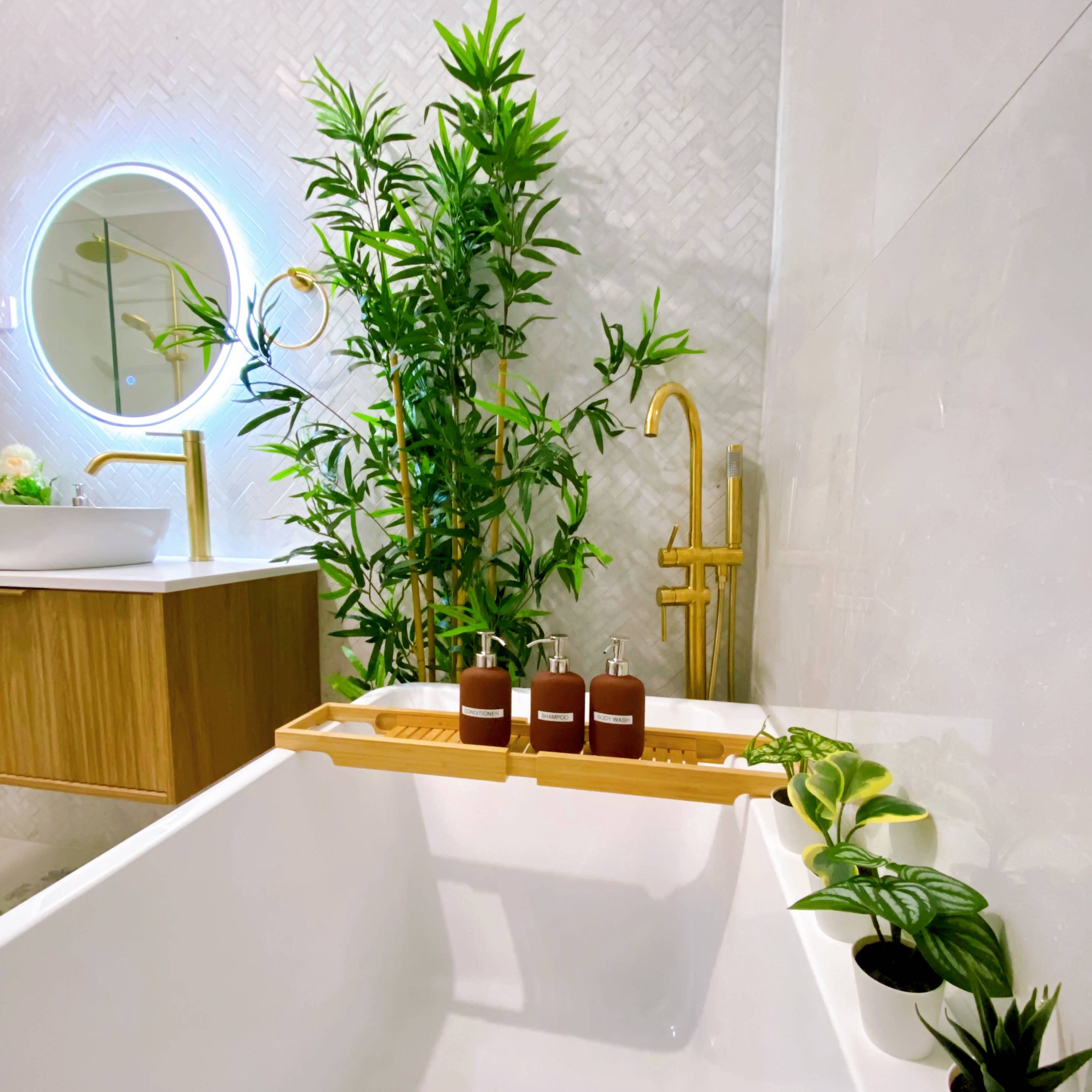 A modern bathroom features a white bathtub with a wood tray holding three bottles, surrounded by potted plants and gold fixtures.