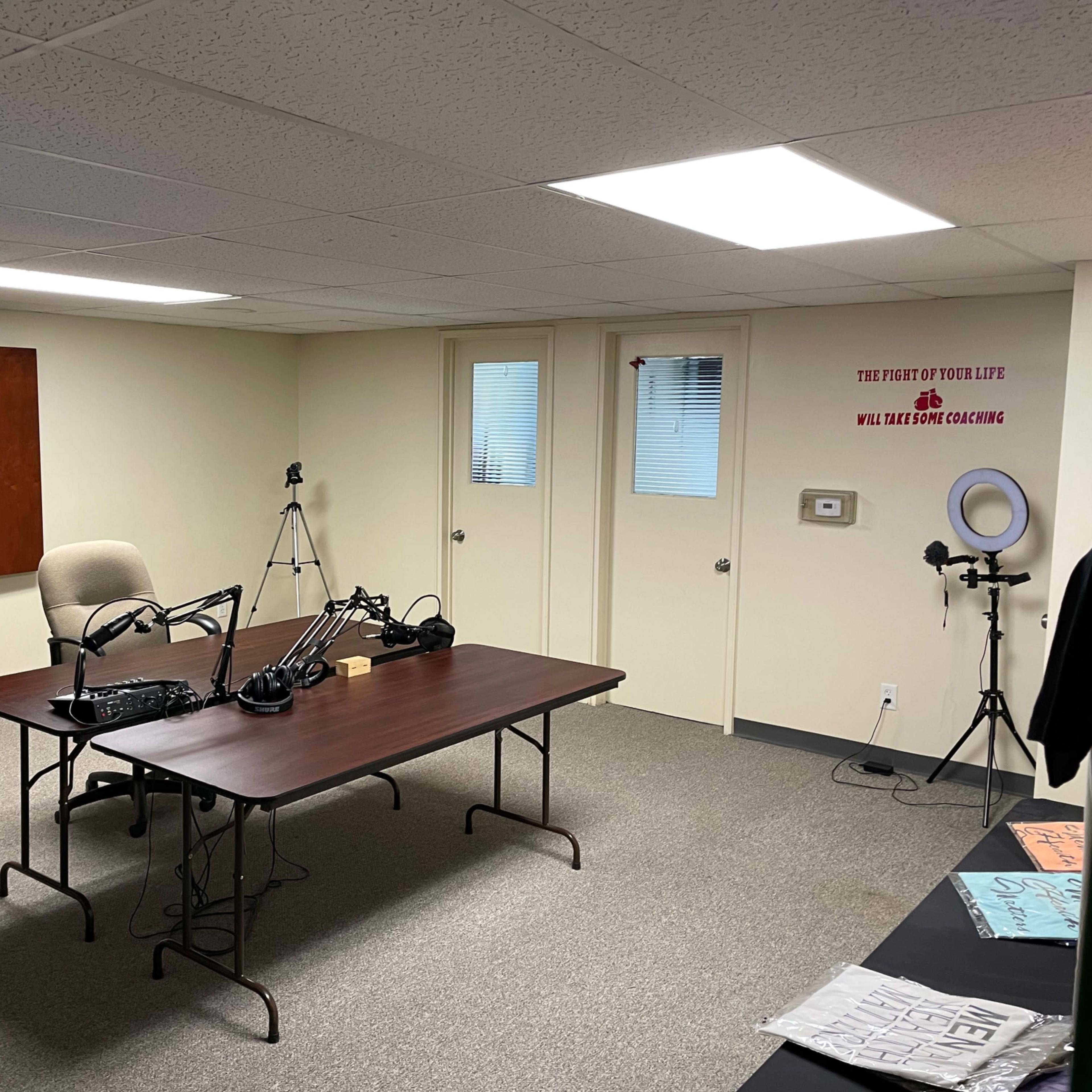 A recording studio with a large table, microphone setups, and a tripod in a sparsely furnished room.