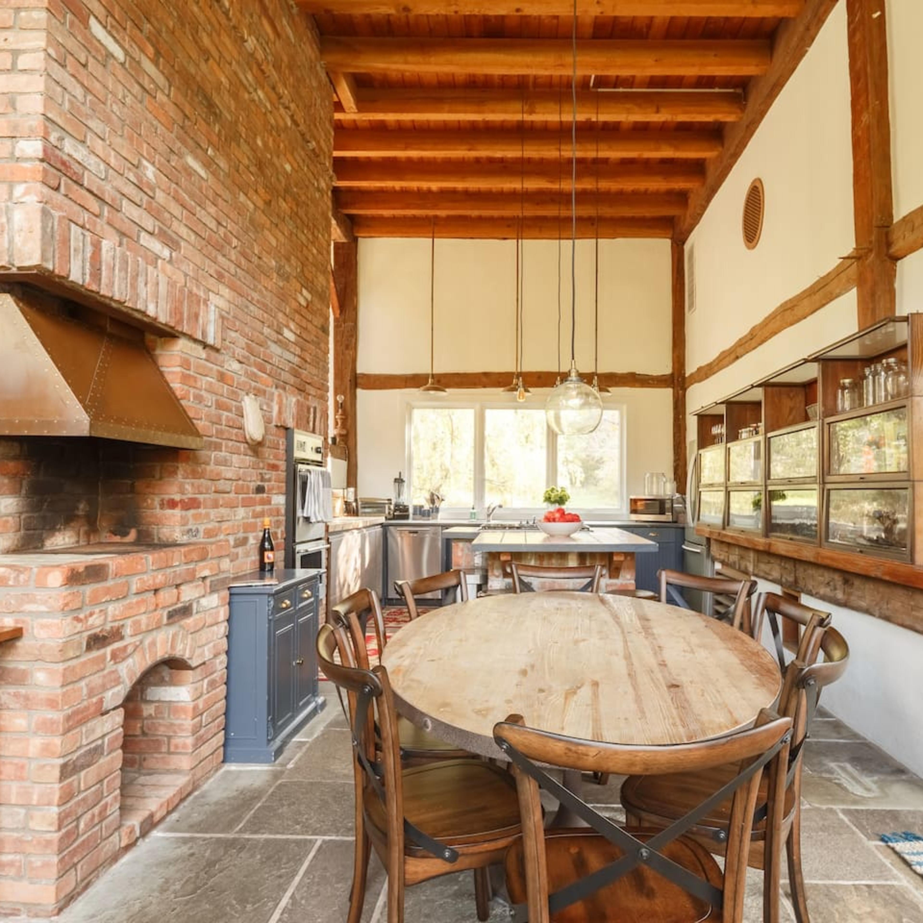 A spacious kitchen with a large wooden dining table, exposed brick walls, a high ceiling with wooden beams, and a modern kitchen setup in the background.