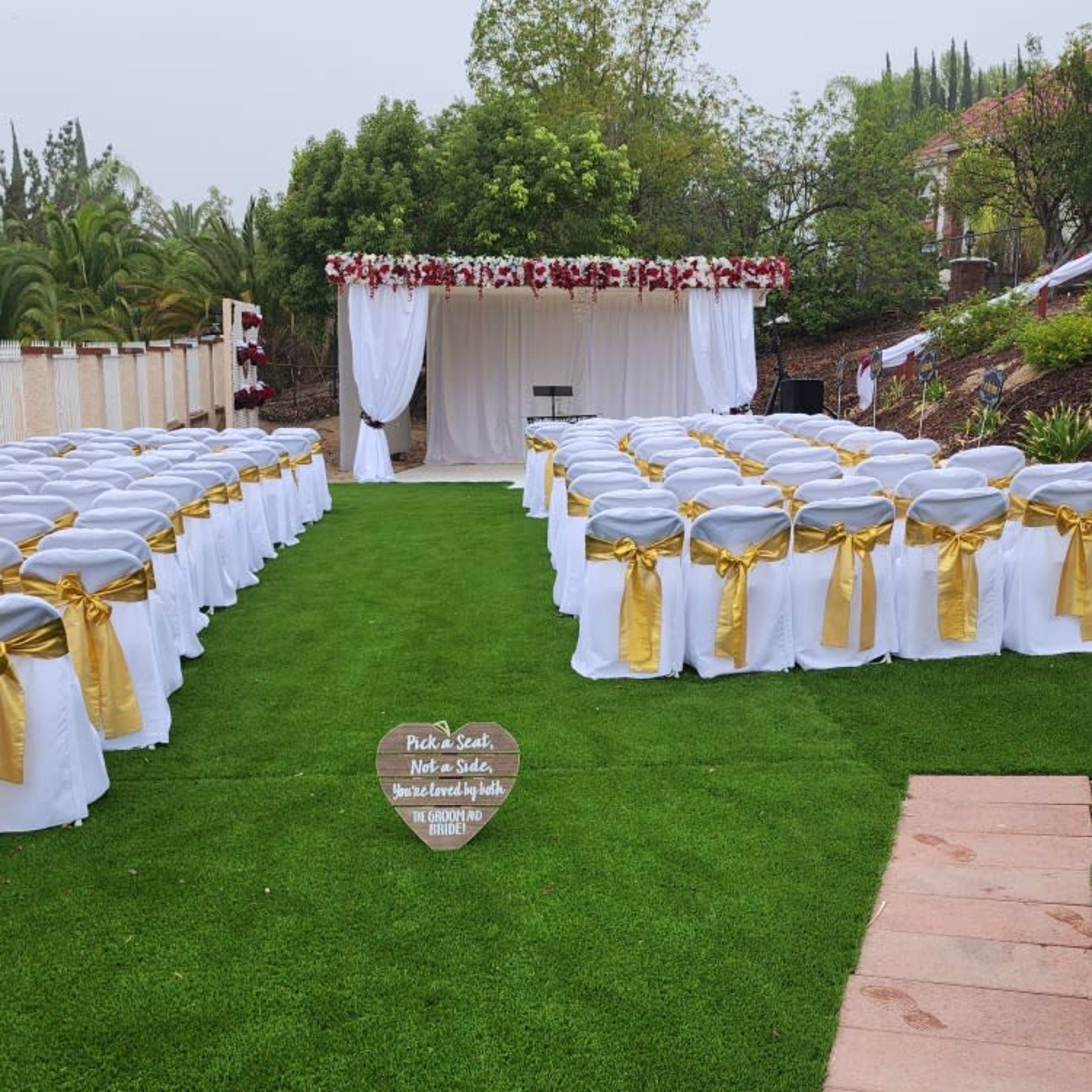 A wedding ceremony setup features rows of white chairs adorned with gold ribbons facing a decorated altar under a canopy.