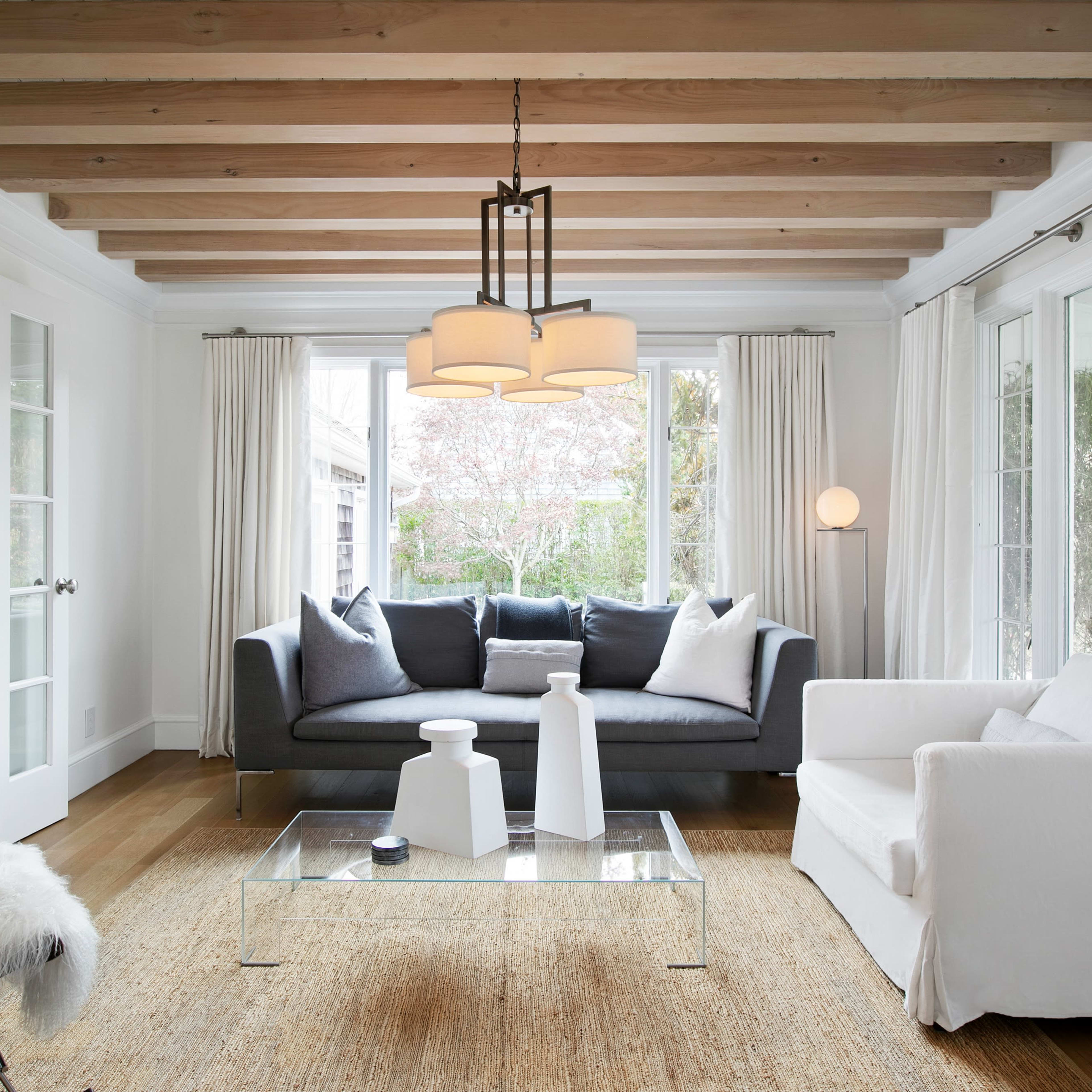 The living room features a gray sofa and a white chair set against a backdrop of large windows and wooden beams overhead.