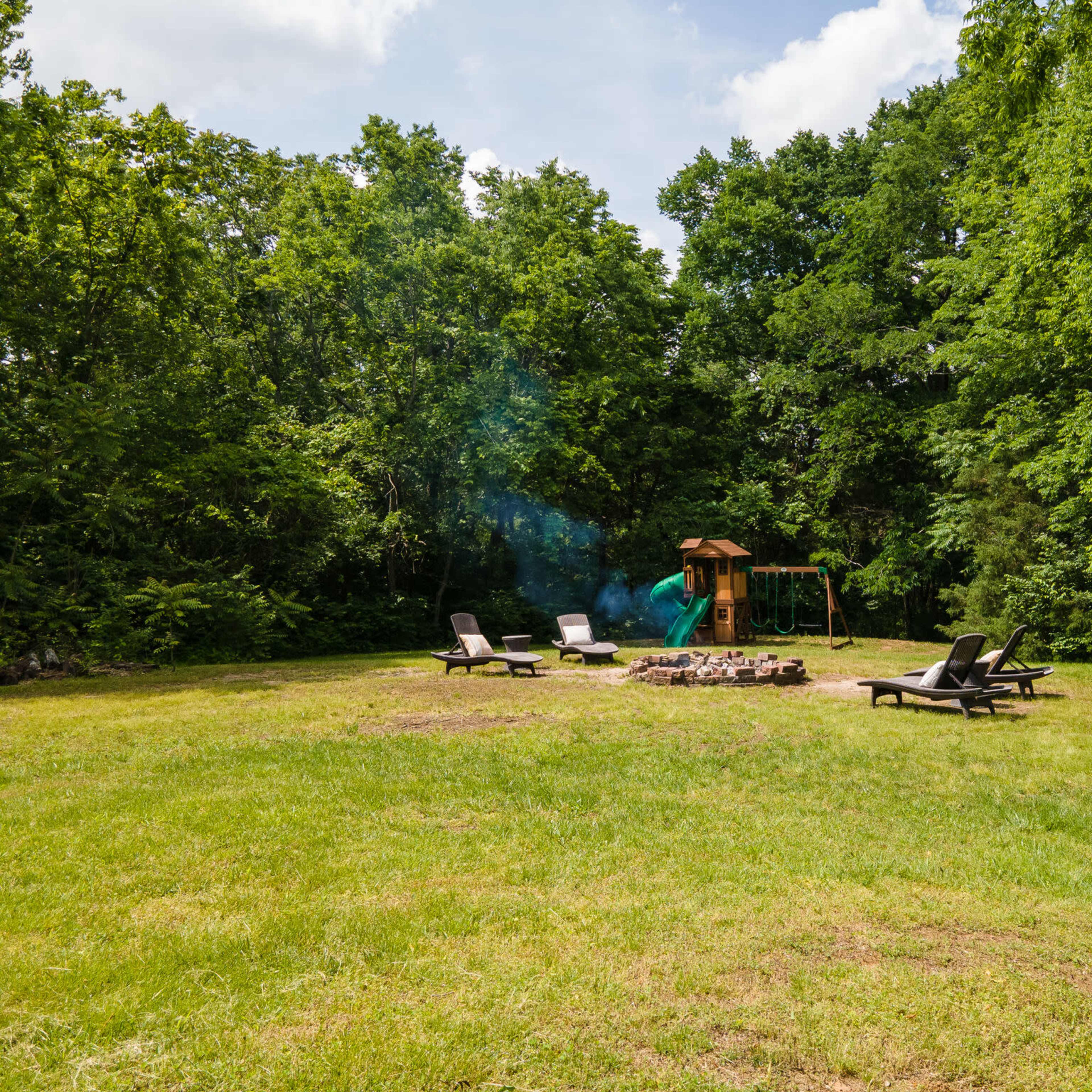 A grassy area surrounded by trees, featuring lounge chairs arranged around a fire pit and a playground structure in the background.