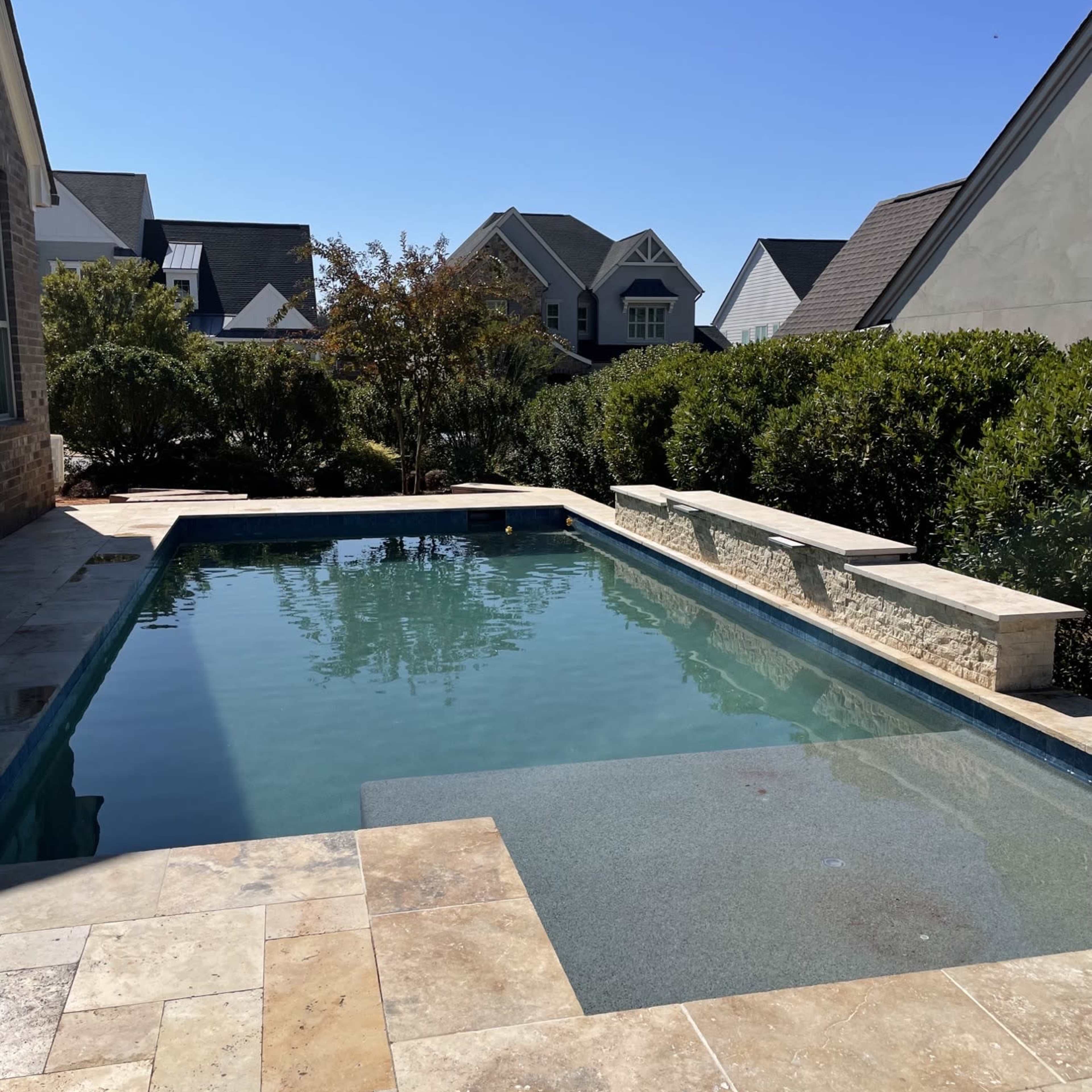 A rectangular swimming pool surrounded by stone tiles and greenery, set against a backdrop of houses under a clear blue sky.