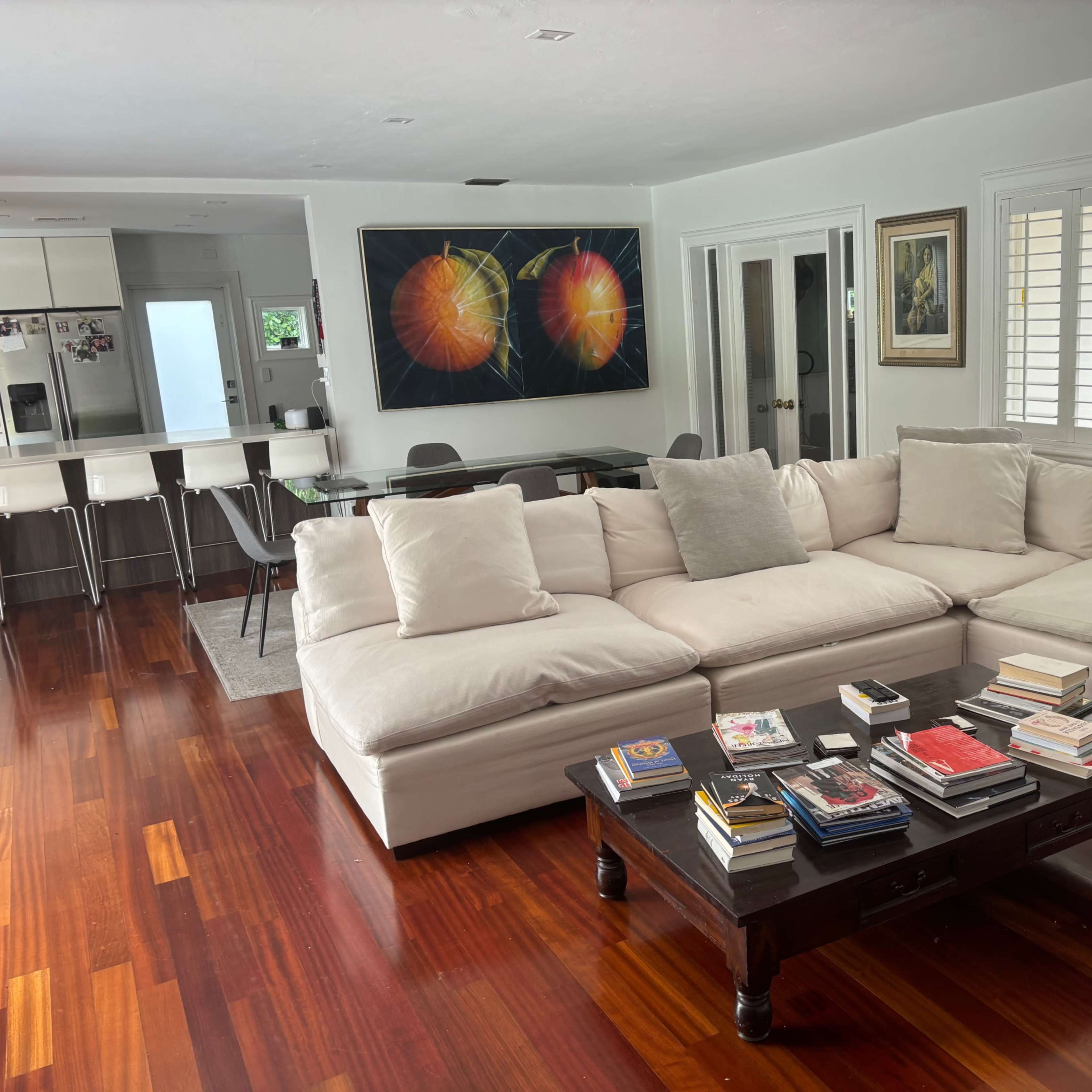 A sectional sofa with white cushions is arranged in a living room next to a coffee table stacked with books, with a dining area visible in the background.