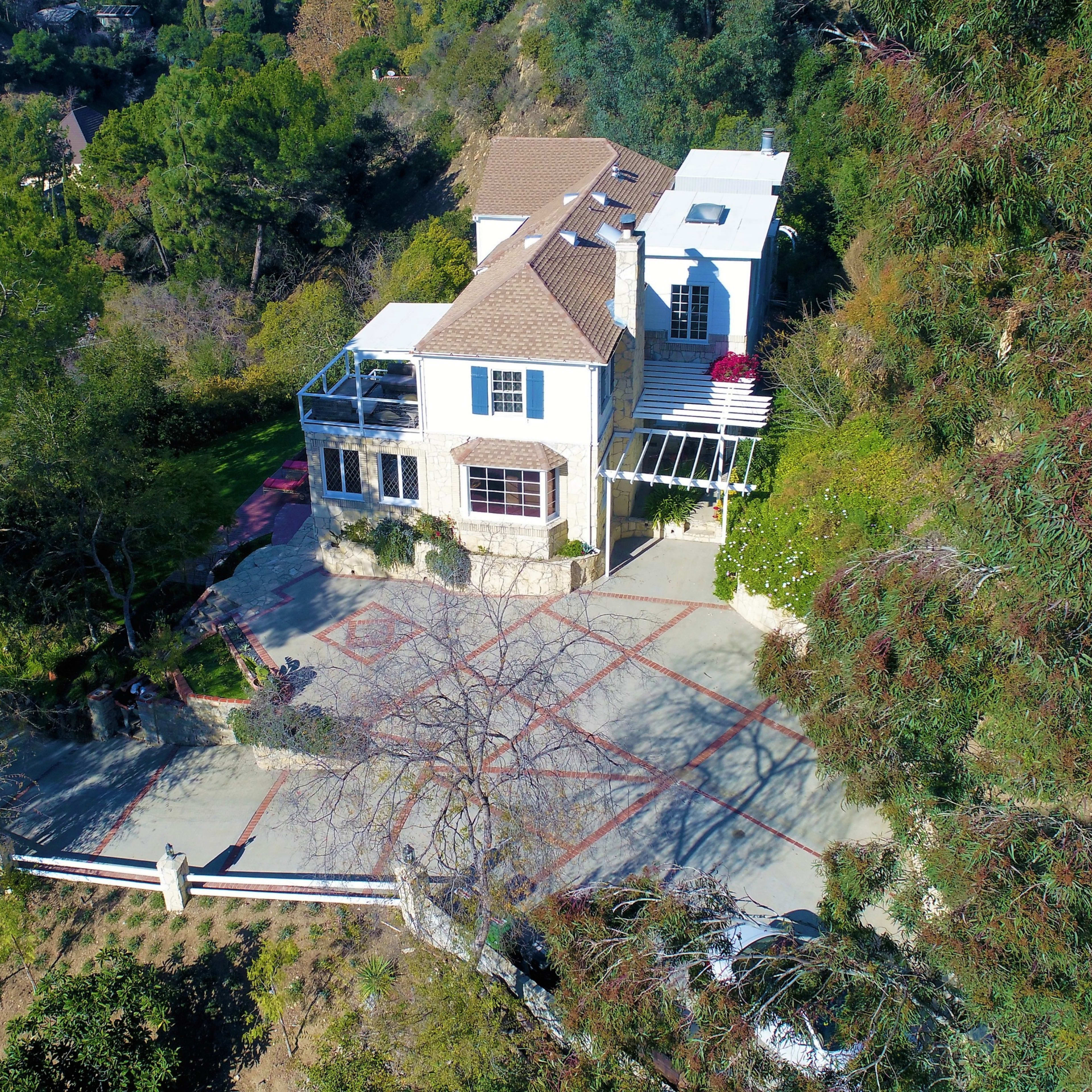 A large, two-story house situated on a hillside surrounded by greenery and a paved driveway.