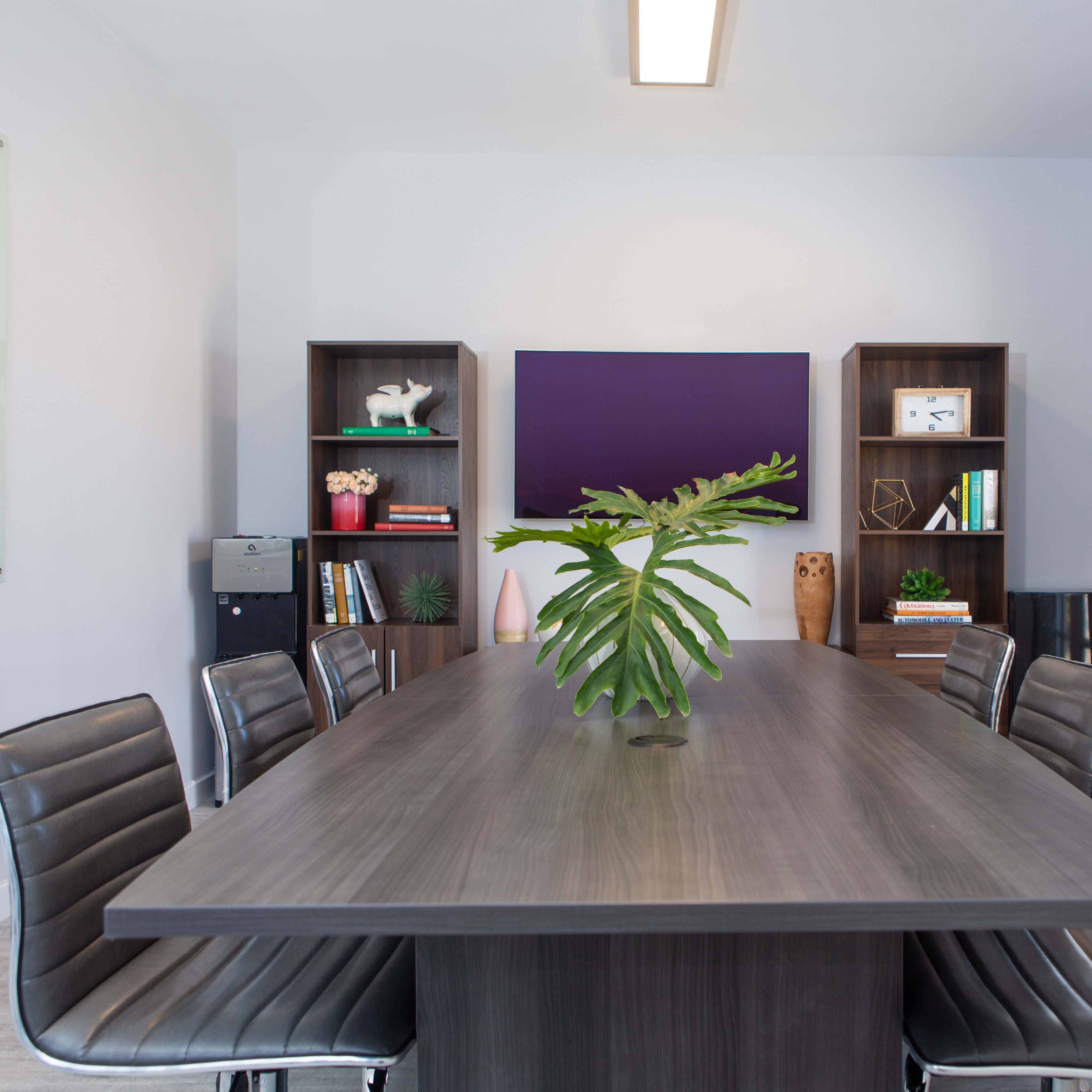 A modern conference room features a long table surrounded by sleek chairs, with a plant centerpiece and shelves displaying decor and books.