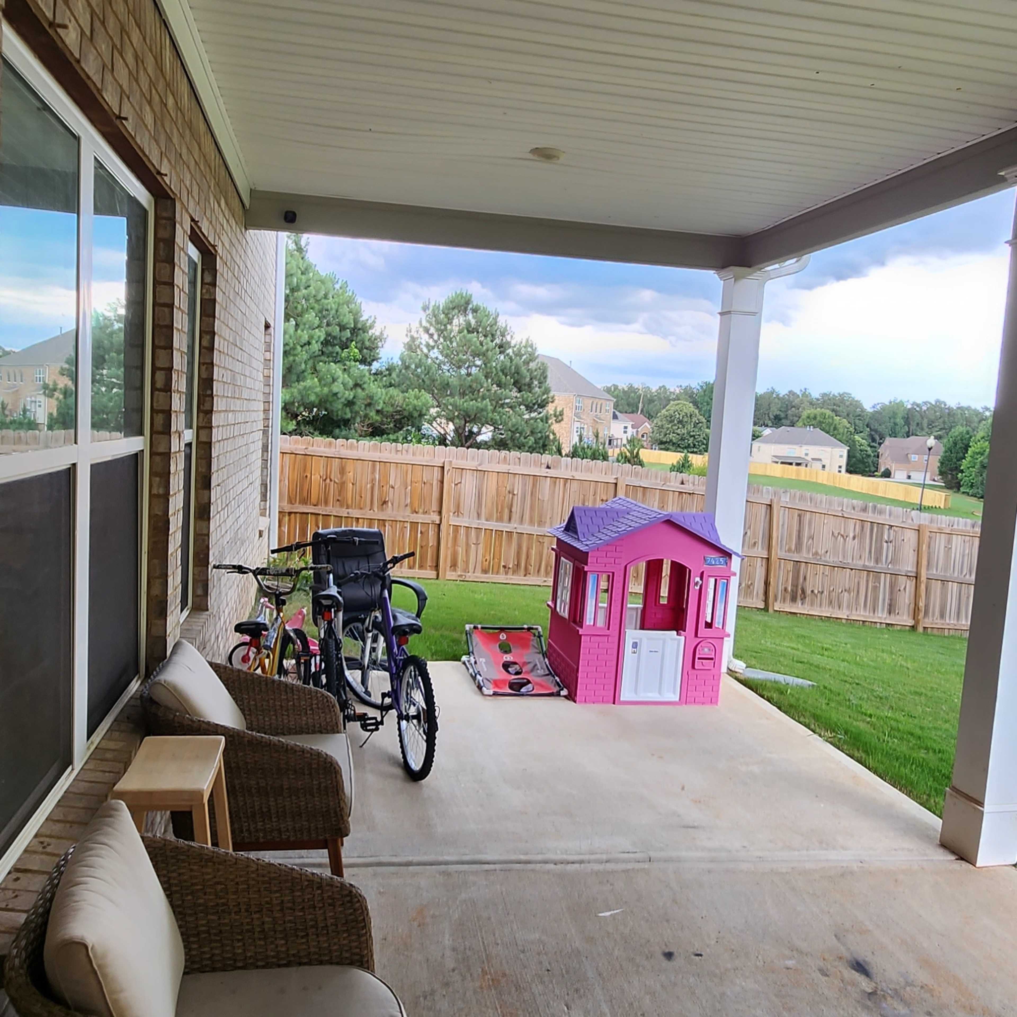 A covered patio features a couple of wicker chairs, a bicycle, and a pink playhouse on a concrete floor.