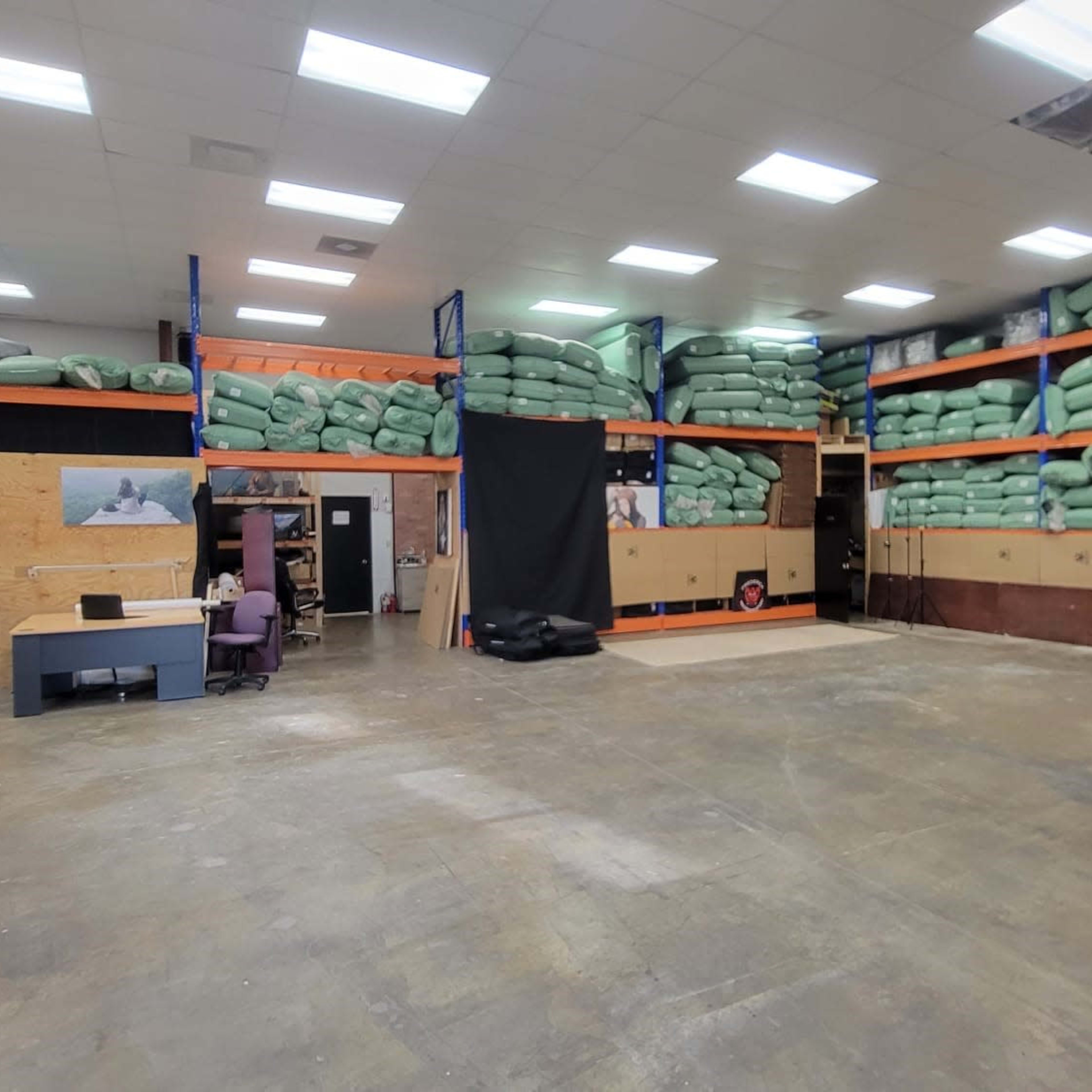 A spacious warehouse interior with stacked green bags on shelves and a workspace featuring a desk and chairs alongside a black backdrop.
