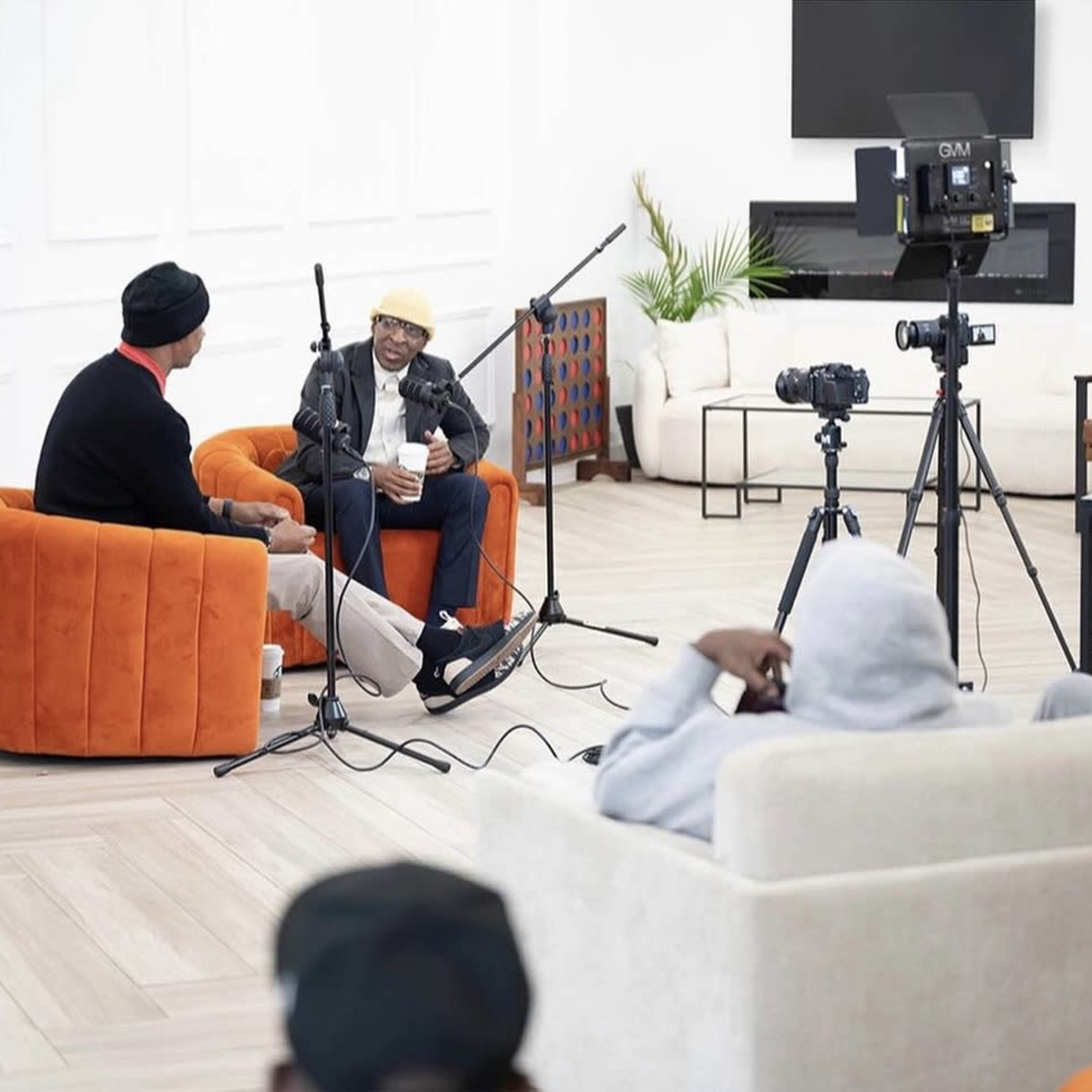 Two men are seated in an orange armchair and a black leather chair, engaged in an interview with cameras and microphones positioned around them.